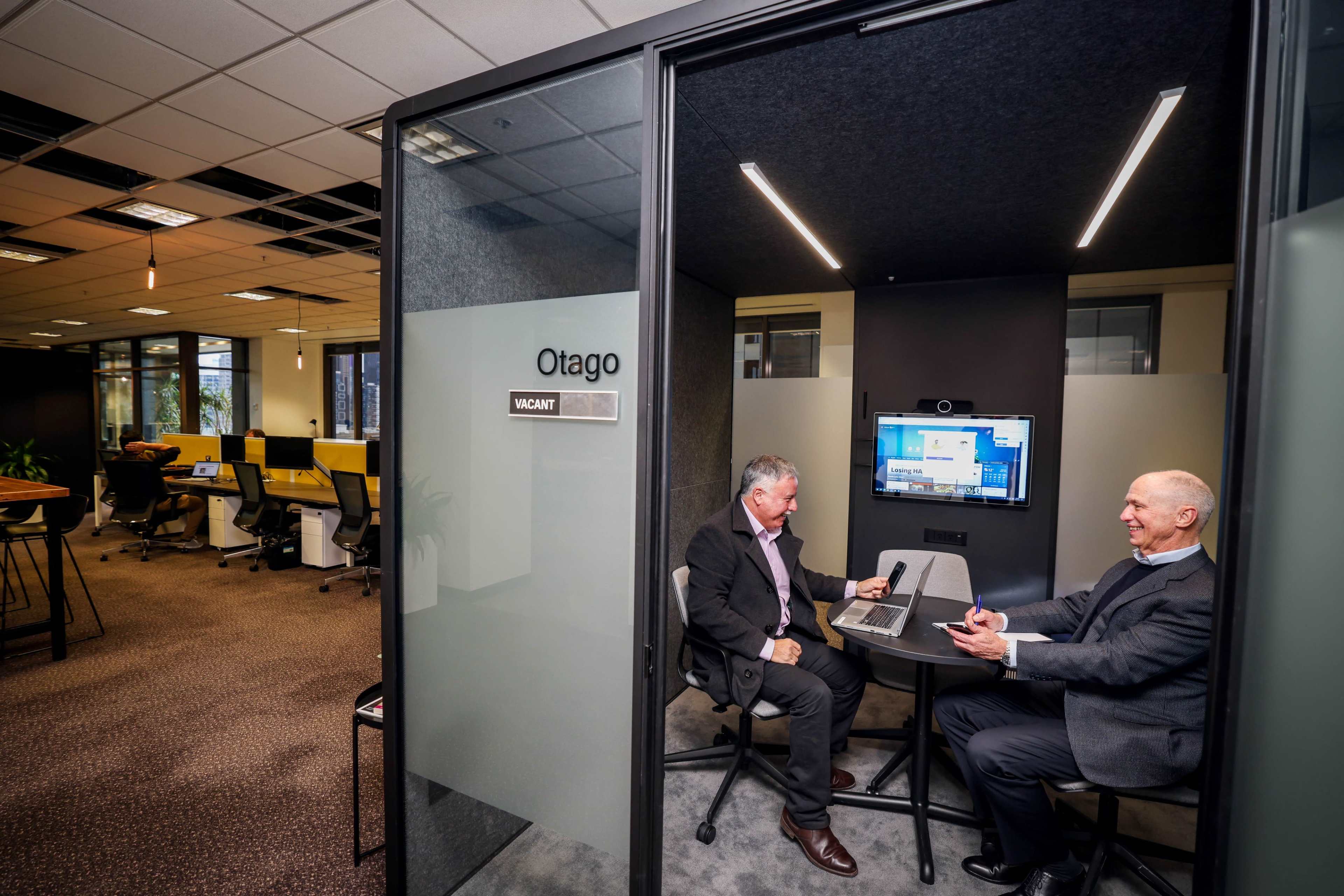 Two men are seated at a round table in a meeting room labeled "Otago," with a screen displaying a presentation in the background.