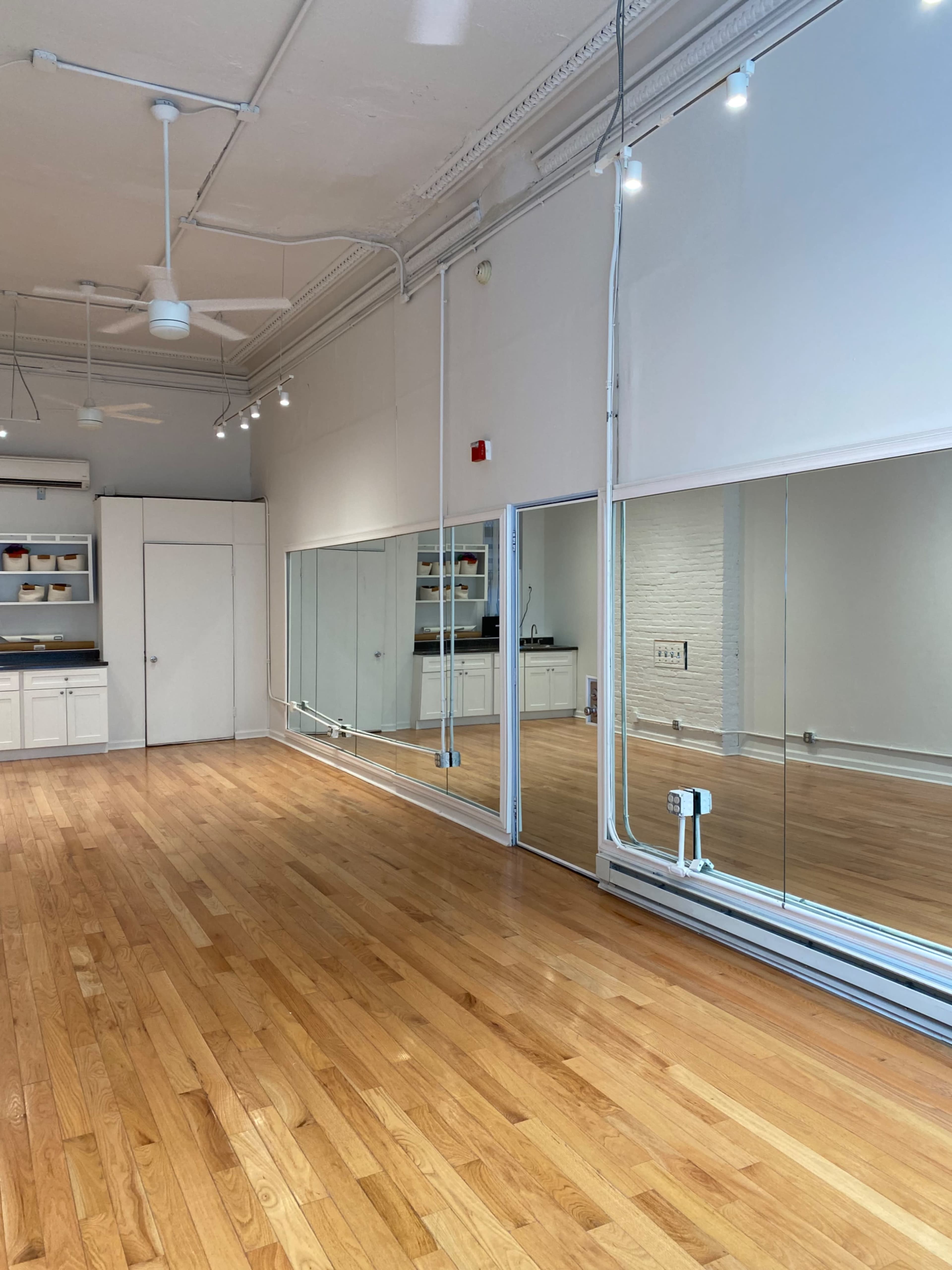 A spacious room with wooden floors, large mirrors along one wall, and a kitchen area in the background.