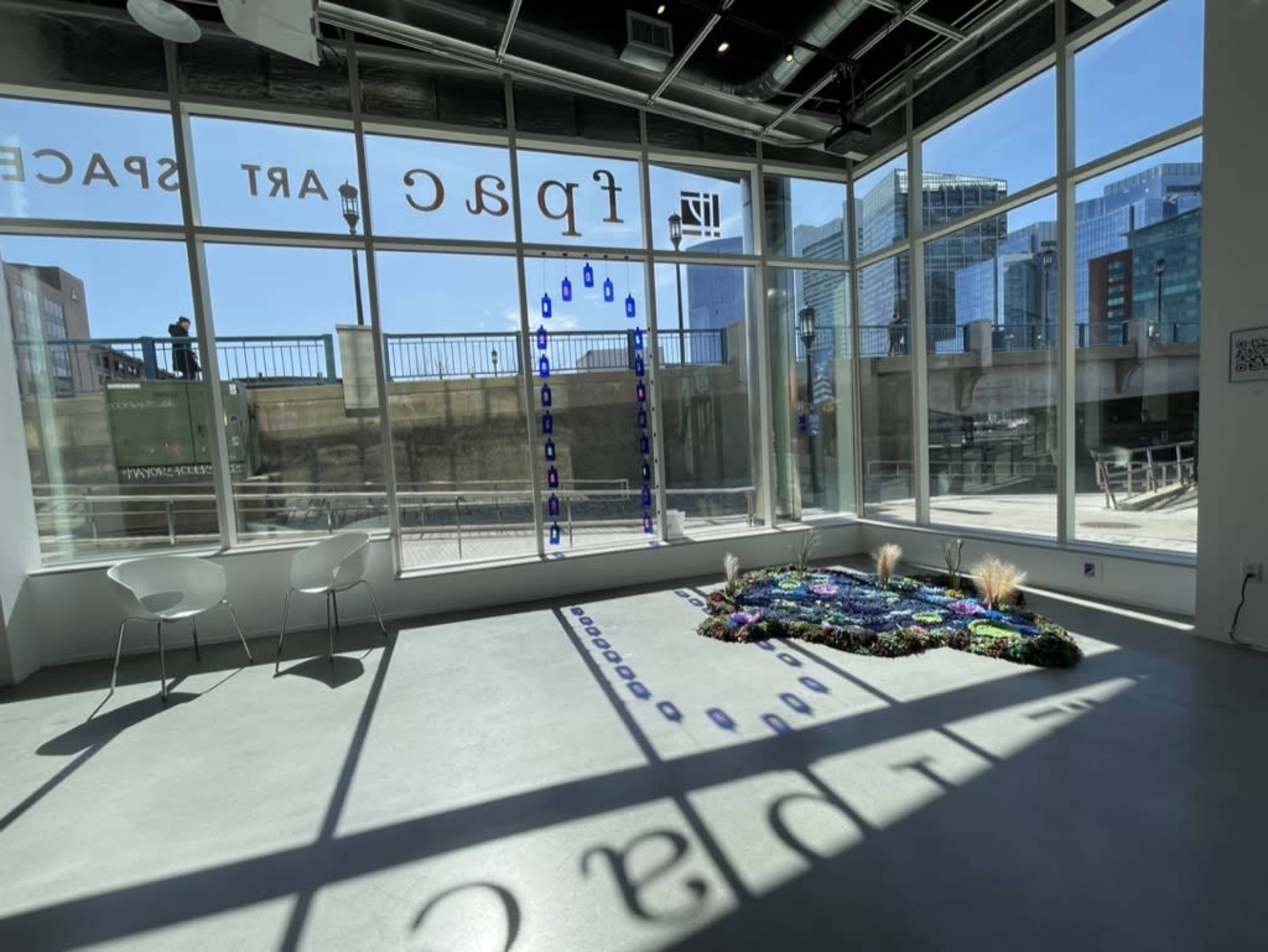 The image shows an art gallery with large windows allowing natural light to illuminate a floor exhibit composed of various colorful materials.