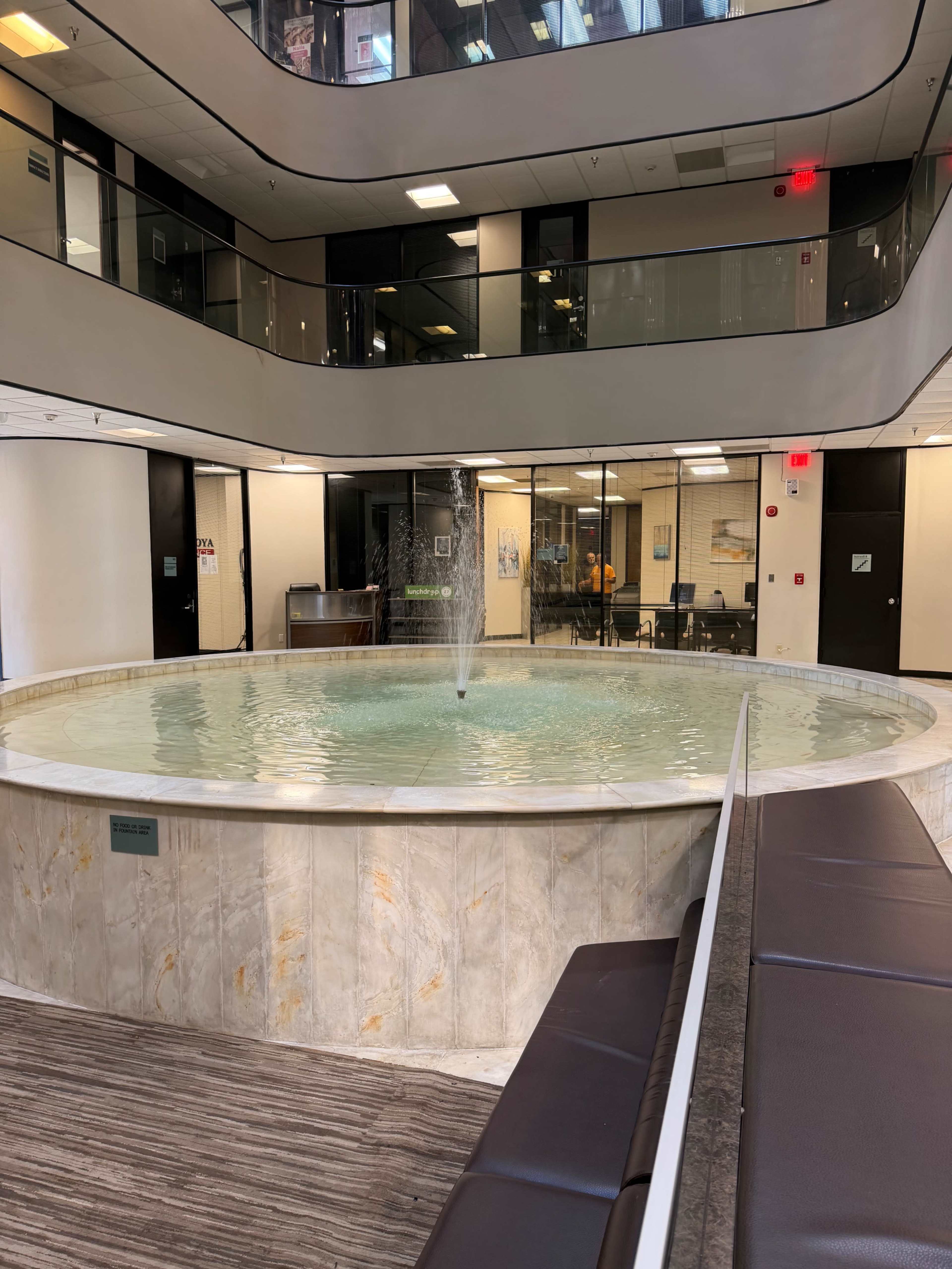 The image shows a circular fountain in the center of a modern indoor space with multiple floors and surrounding seating areas.