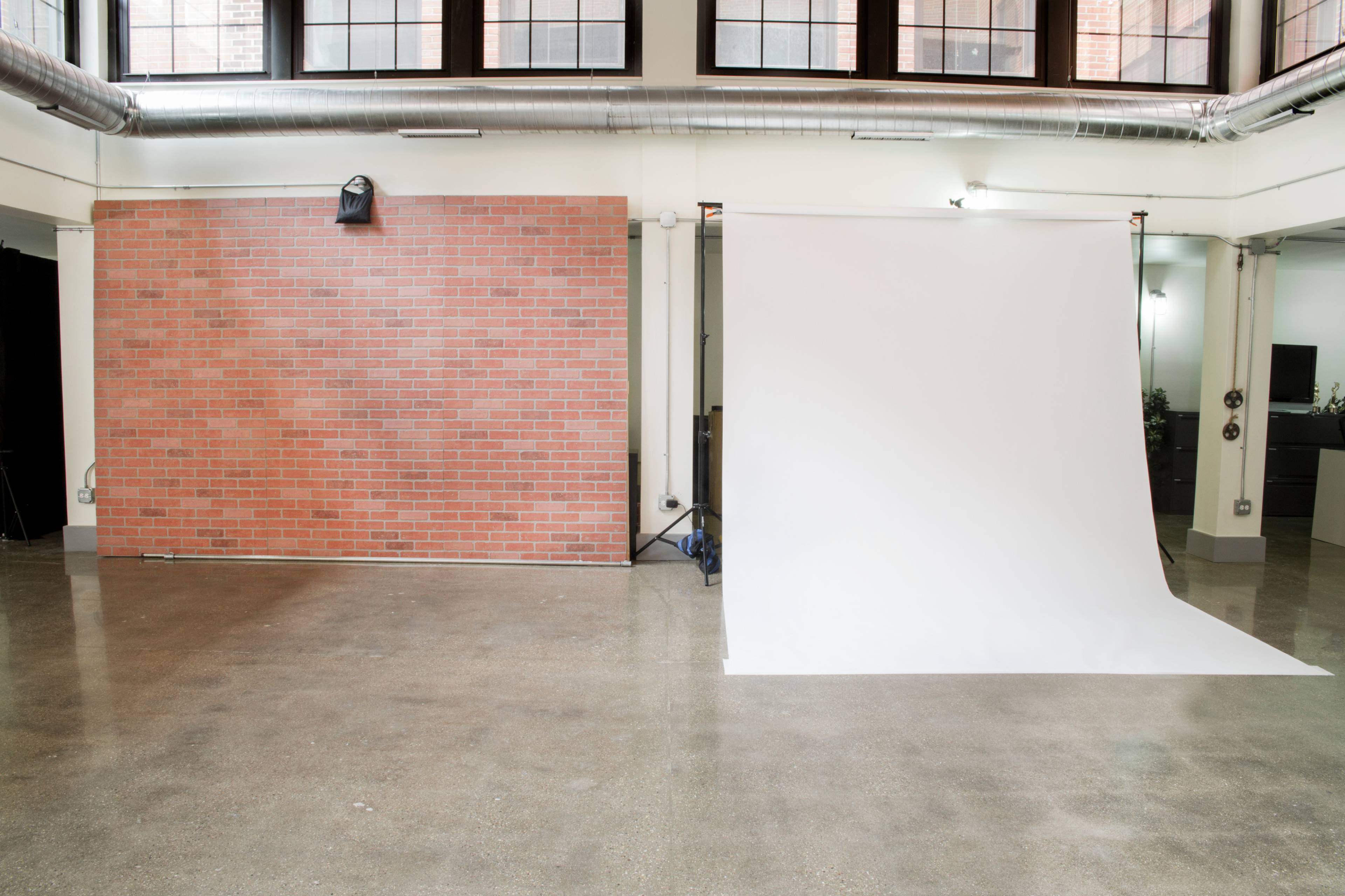 The image shows a spacious studio with a brick wall on one side and a white backdrop on the other, separated by a polished concrete floor.