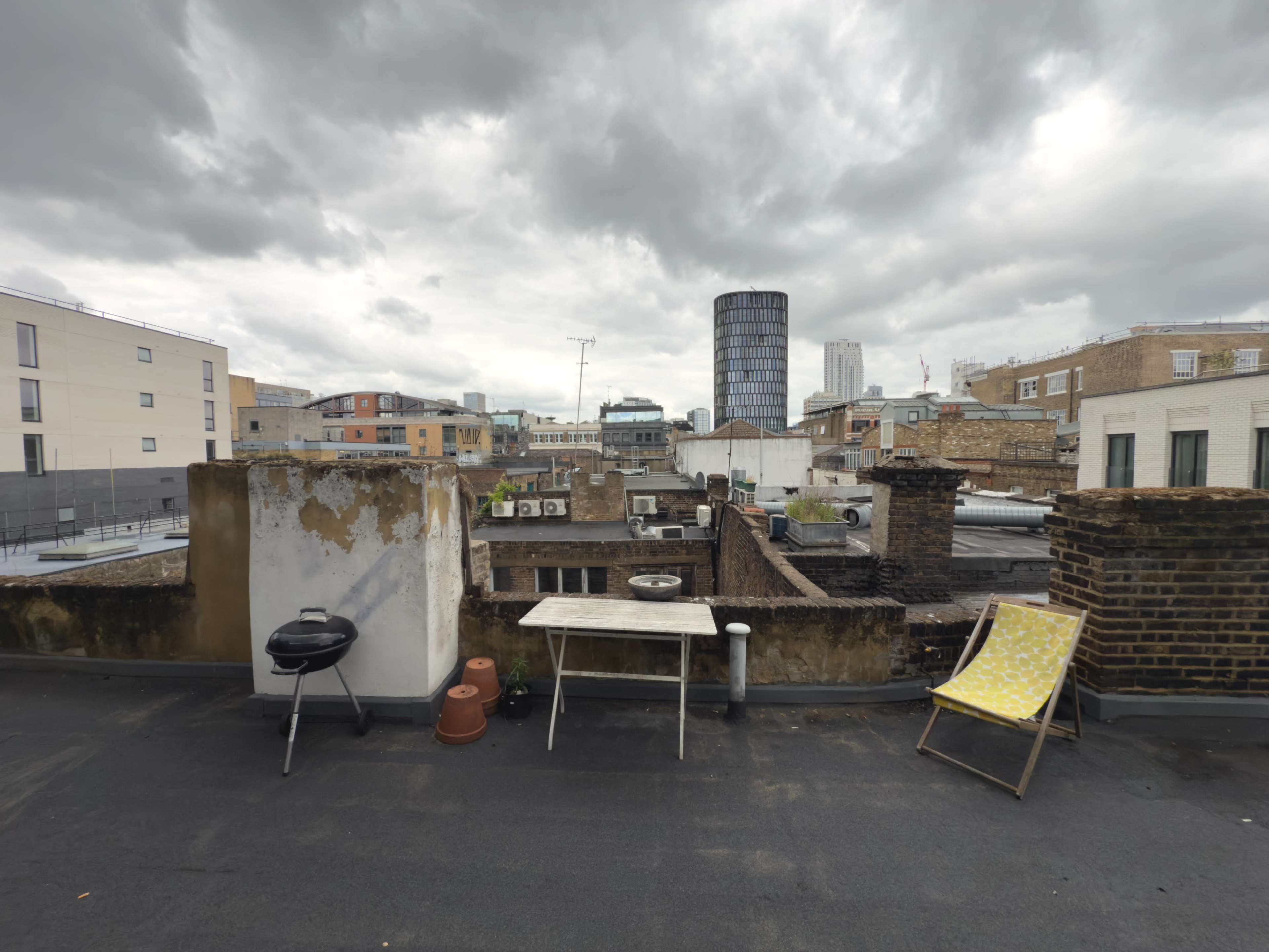 The image shows a rooftop scene with a charcoal grill, a table, a lounge chair, and buildings under a cloudy sky.