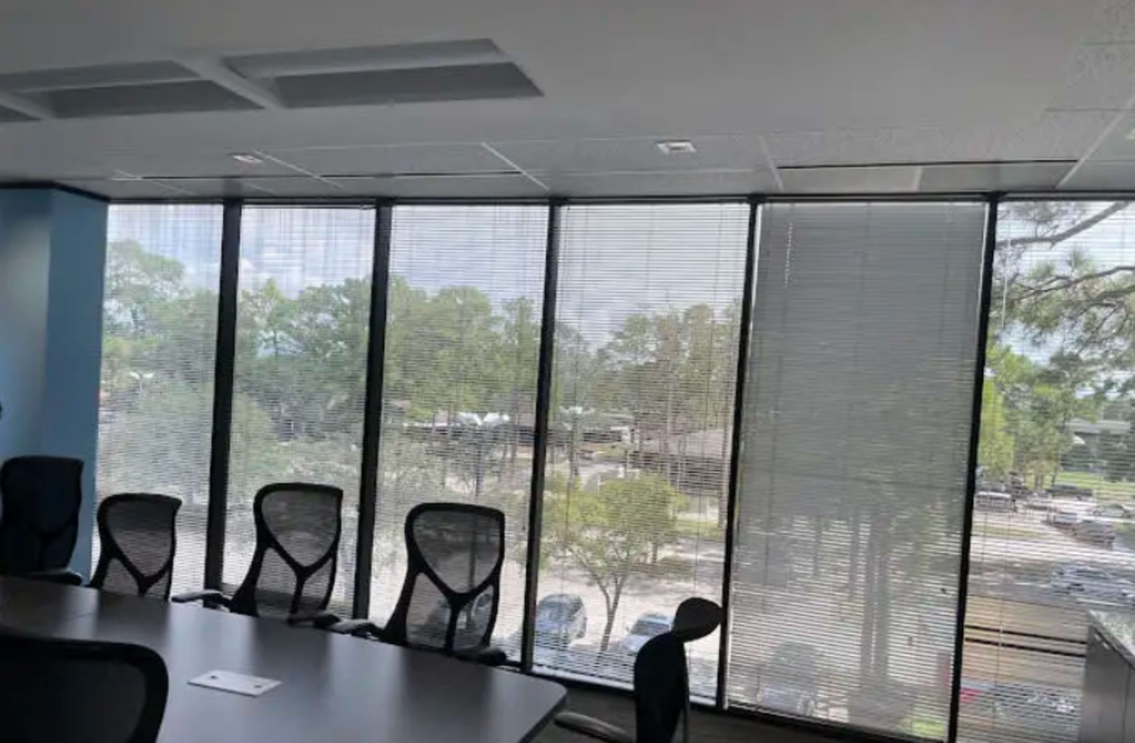 A conference room features a large table surrounded by ergonomic chairs, with large windows displaying a view of trees and a parking area outside.