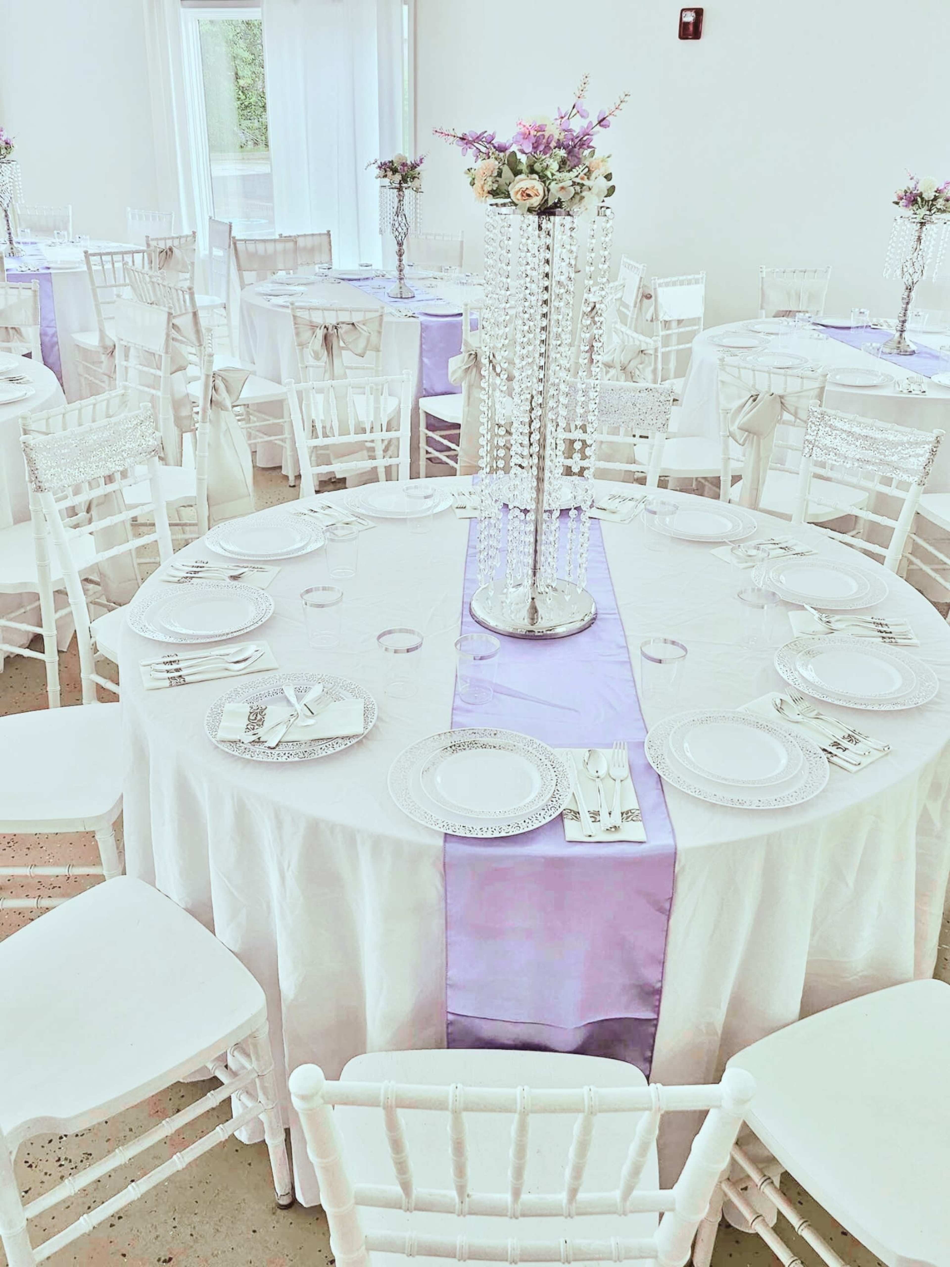 A finely decorated banquet hall features round tables set with white tablecloths, purple runners, and floral centerpieces, surrounded by white chairs.