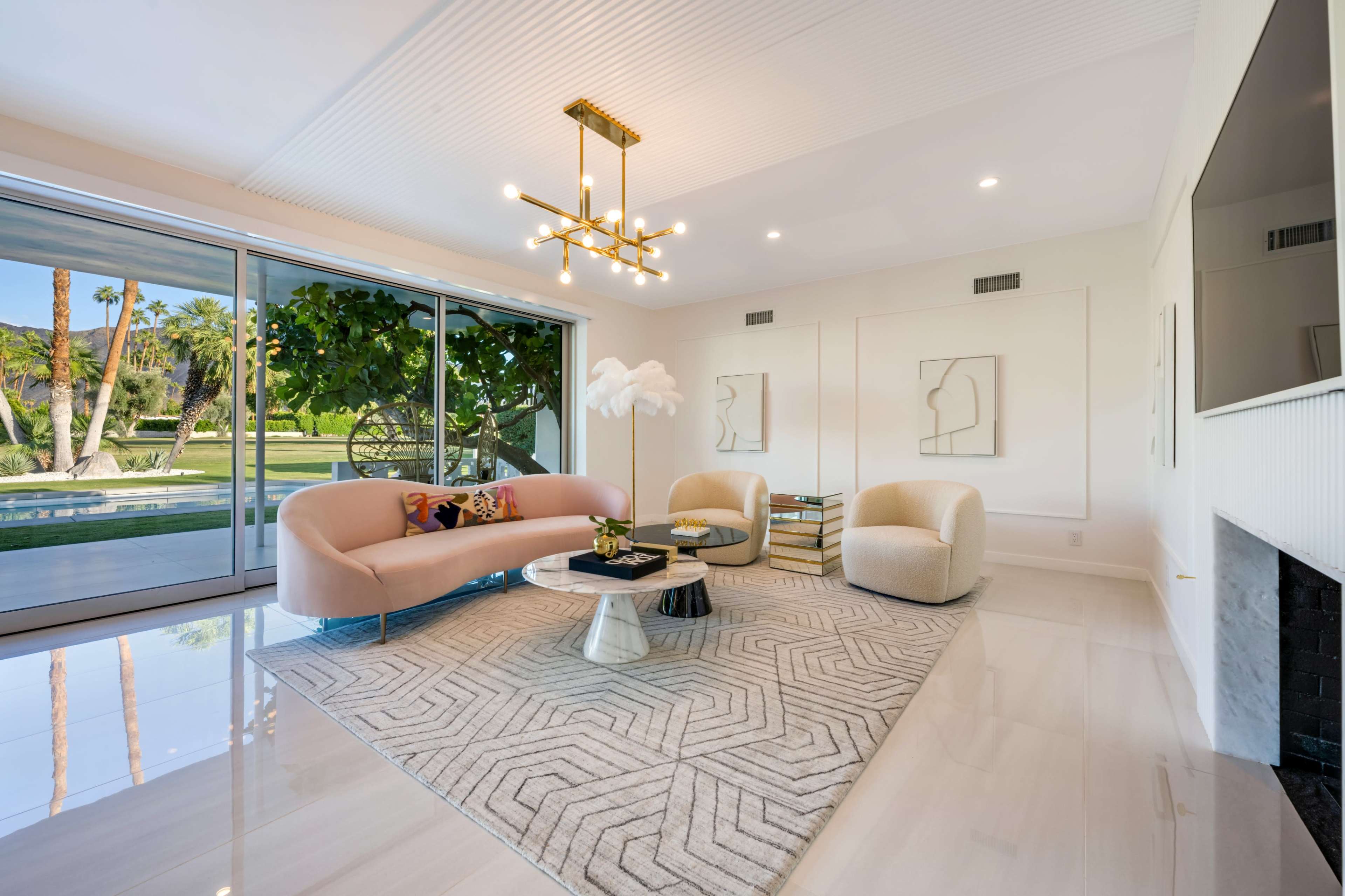 A modern living room features a pink curved sofa, two cream armchairs, a round coffee table on a geometric rug, and large glass doors opening to a garden view.