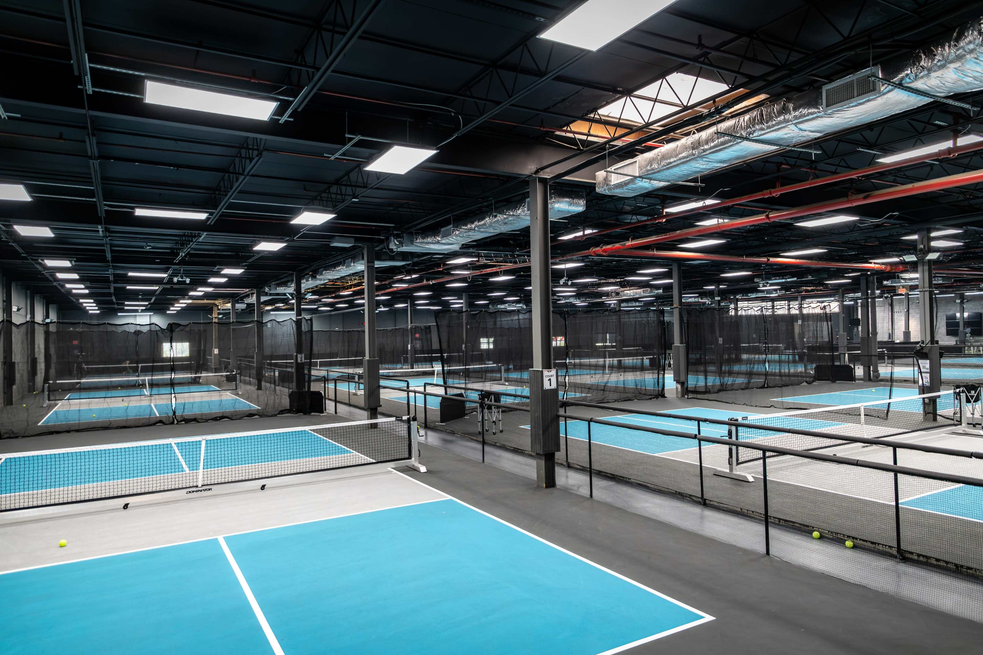 The image shows an indoor sports facility featuring multiple tennis courts with blue surfaces, surrounded by netting and overhead lighting.