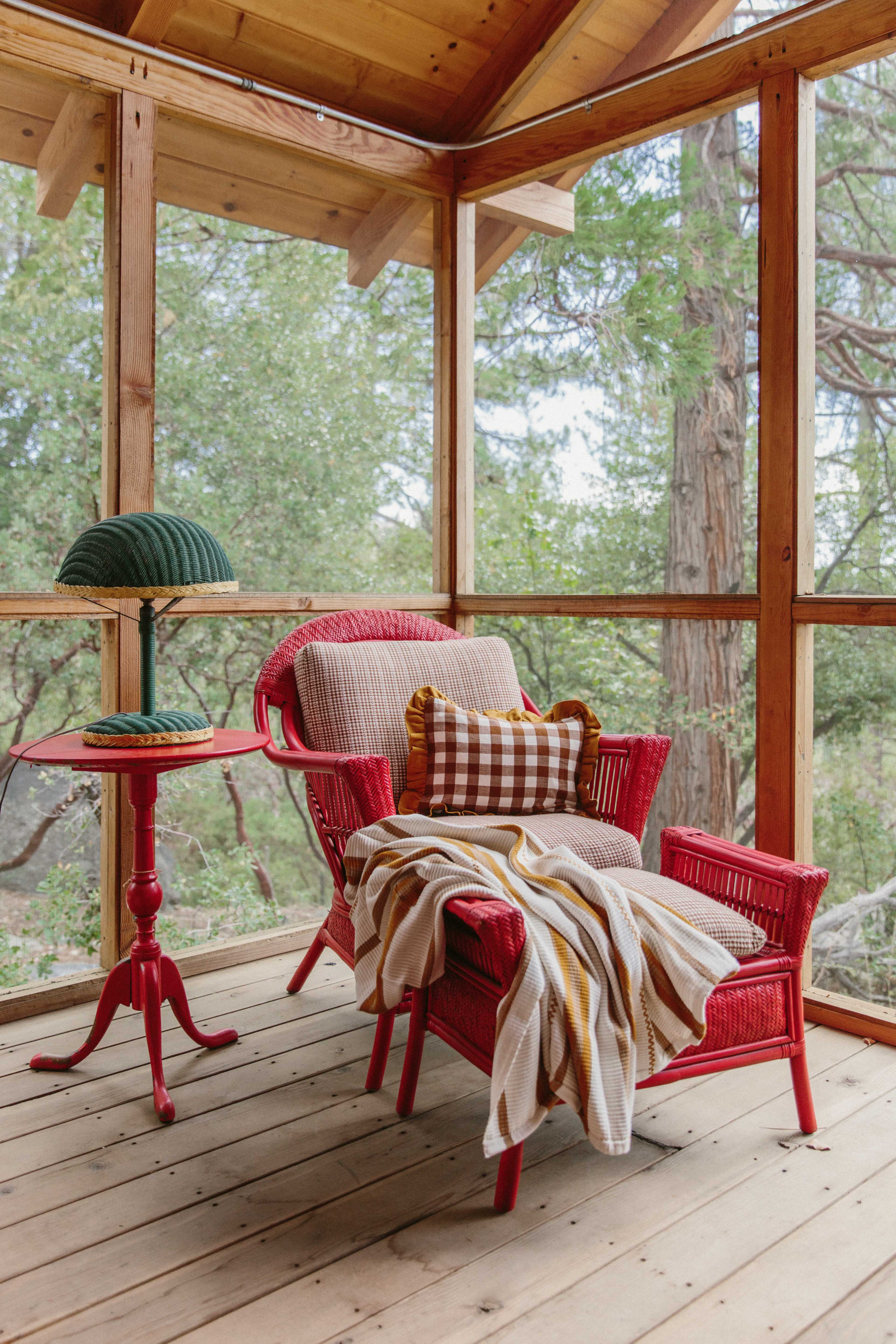 A red wicker chair with a patterned throw blanket and a small side table with a lamp is situated in a screened-in porch surrounded by trees.