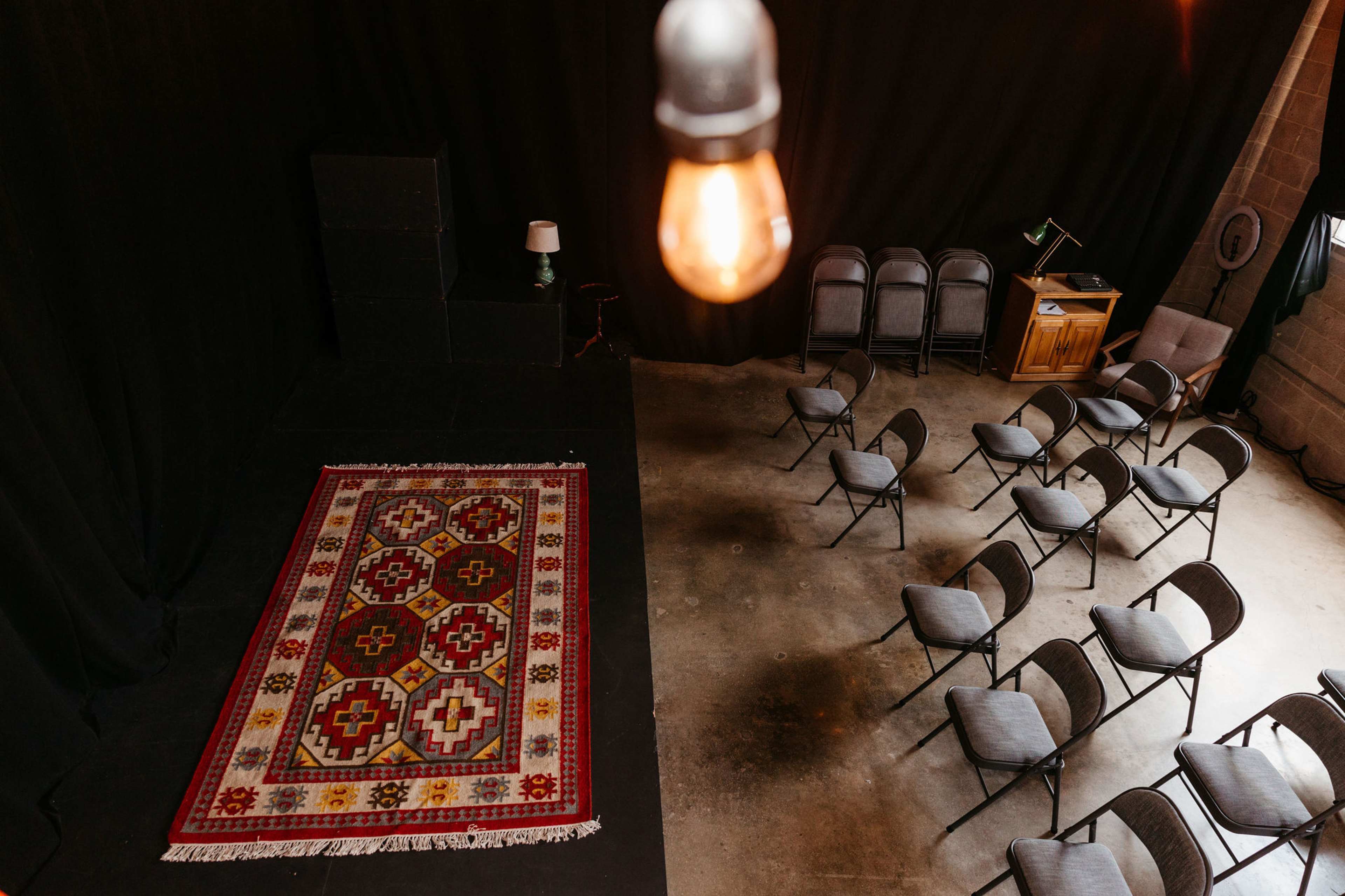 A small stage with a decorative rug, surrounded by empty folding chairs, set in a dimly lit room.