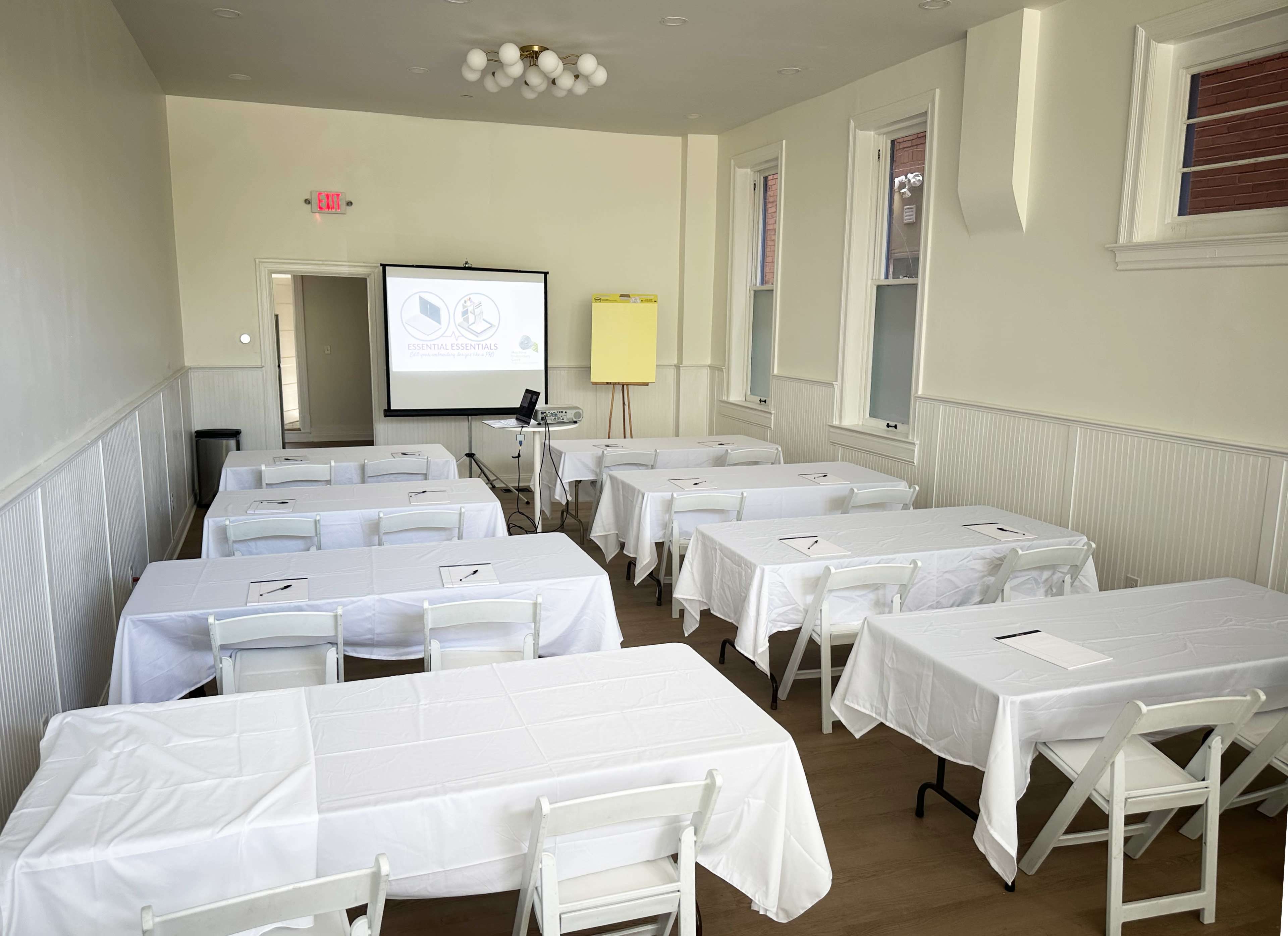 The image shows a meeting room arranged with several tables covered in white tablecloths, facing a presentation screen and a projector setup.