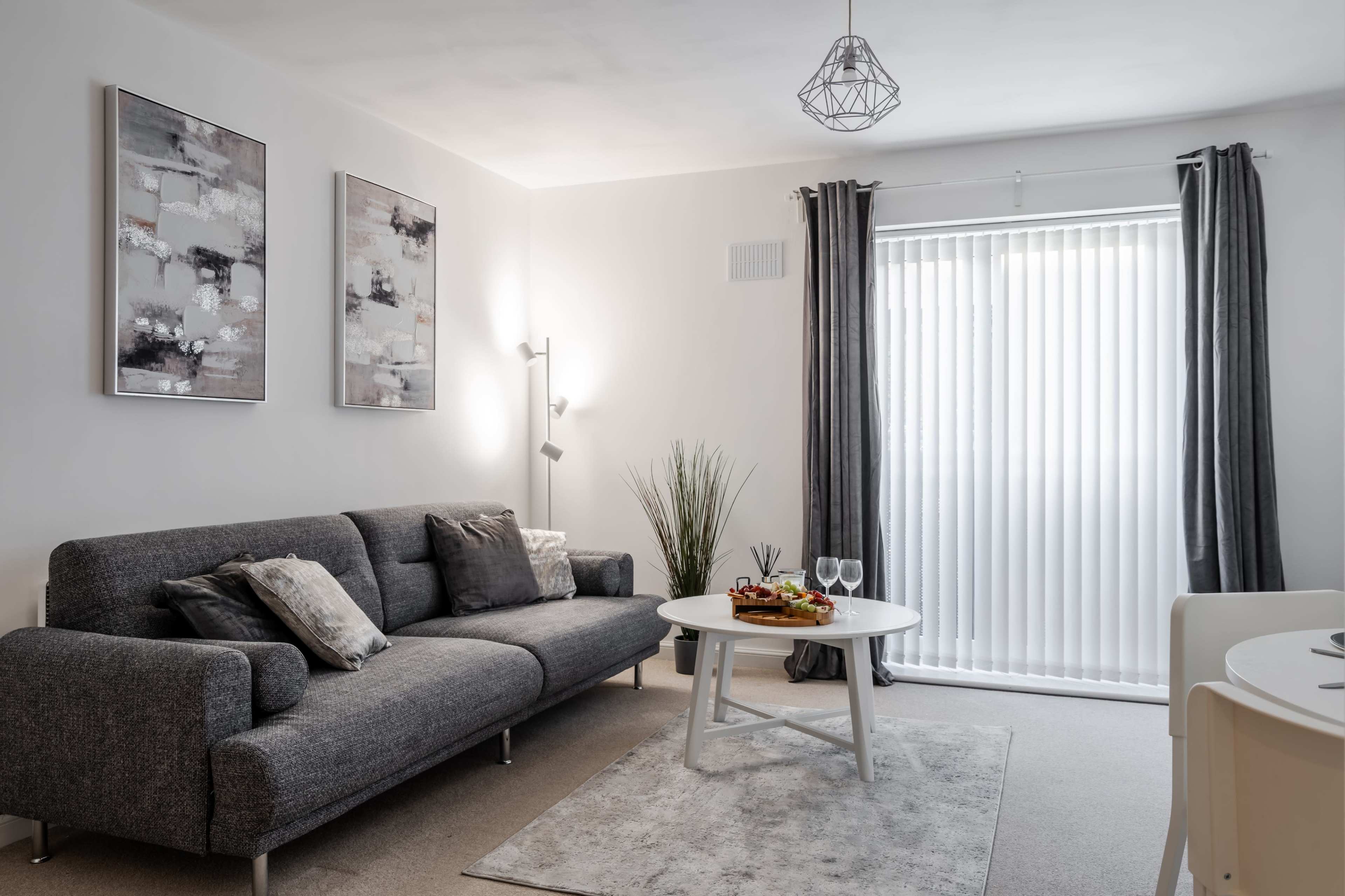 A modern living room features a gray sofa, a white coffee table with snacks, and large windows covered by vertical blinds.