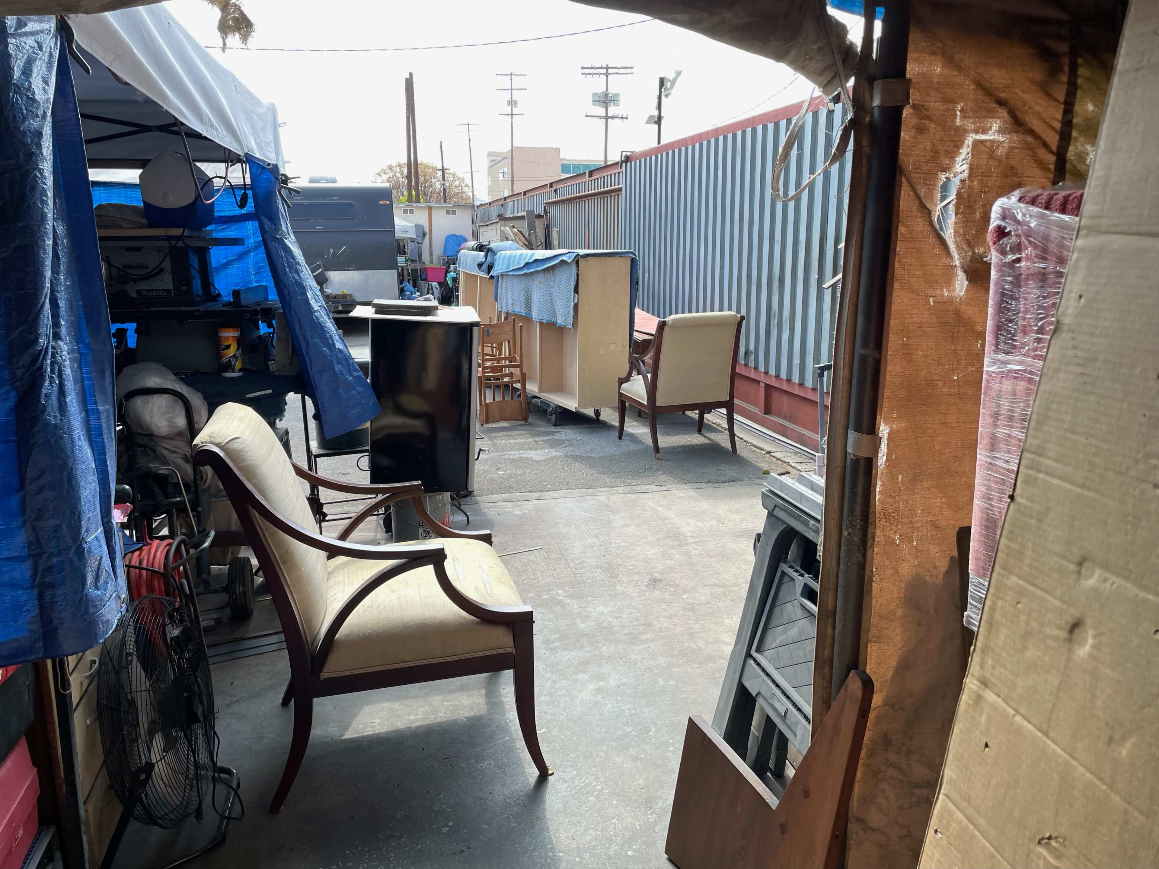 The image shows a narrow alleyway filled with various items, including furniture and tarps, partially enclosed by metal containers and walls.
