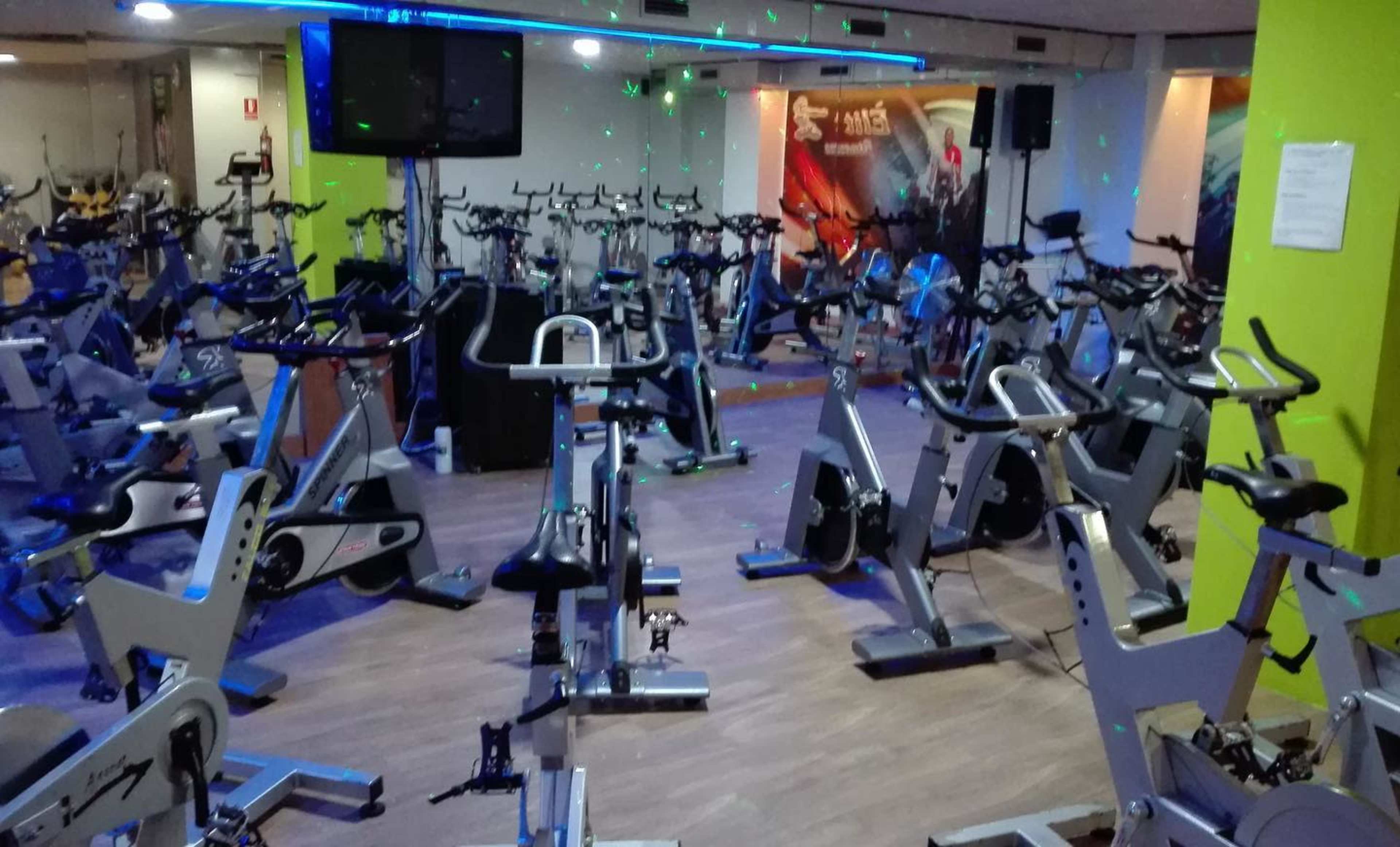 A fitness studio is filled with stationary bicycles and illuminated by colorful lights.