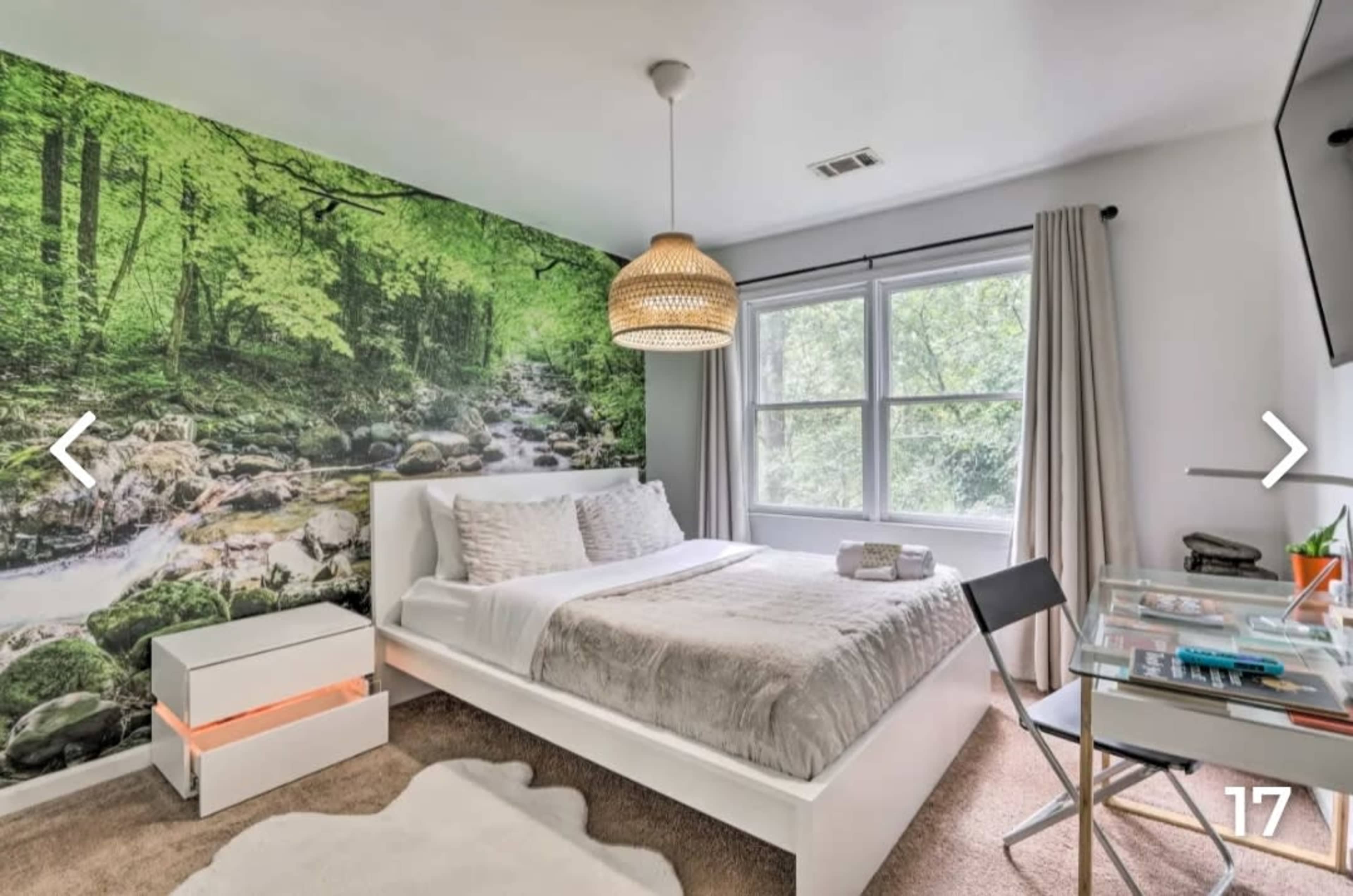 The bedroom features a large mural of a green forest, a white bed with gray bedding, a glass desk, and a light fixture hanging from the ceiling.