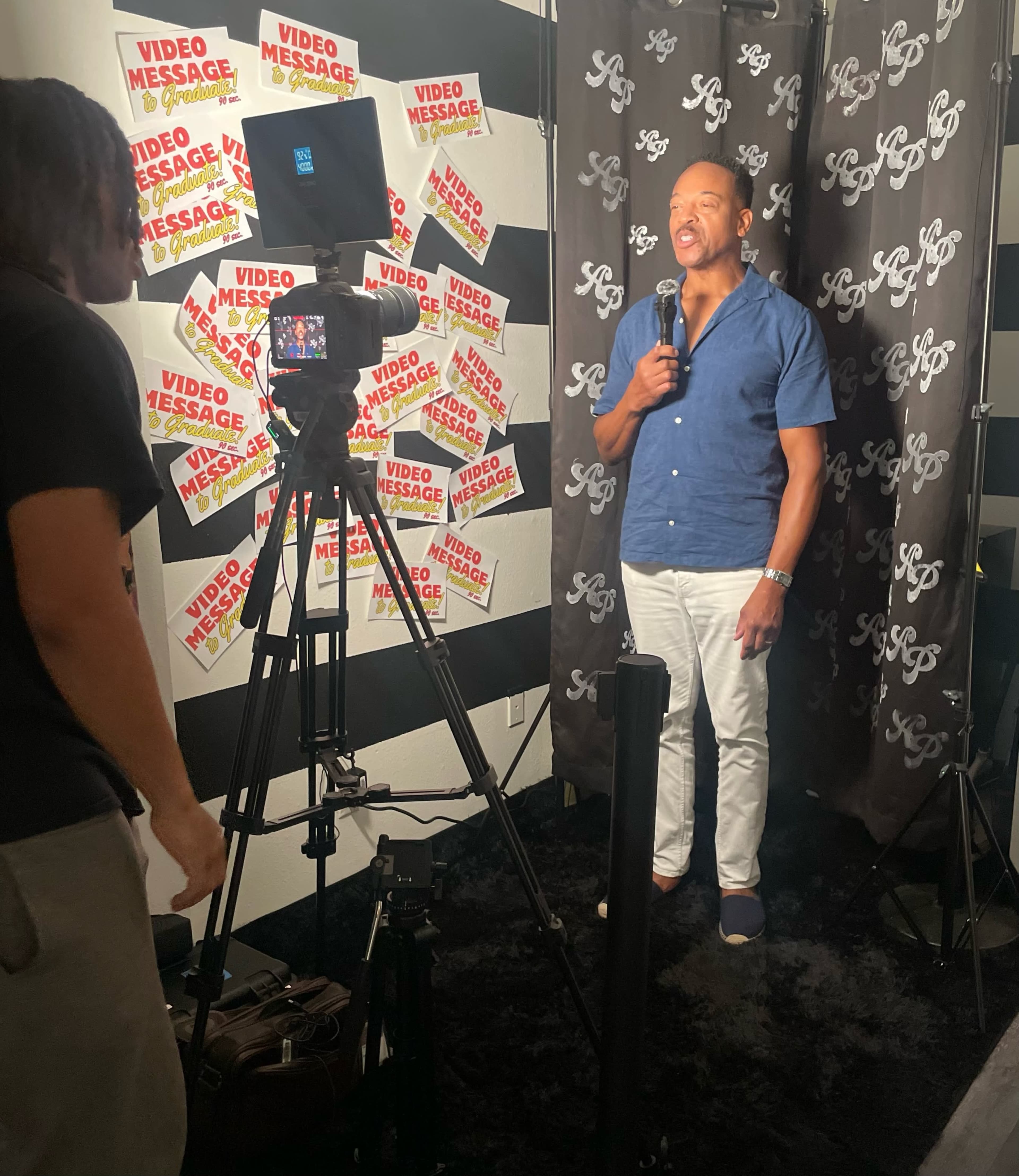 A man stands in front of a video camera, speaking in a studio decorated with signs that read "VIDEO MESSAGE."