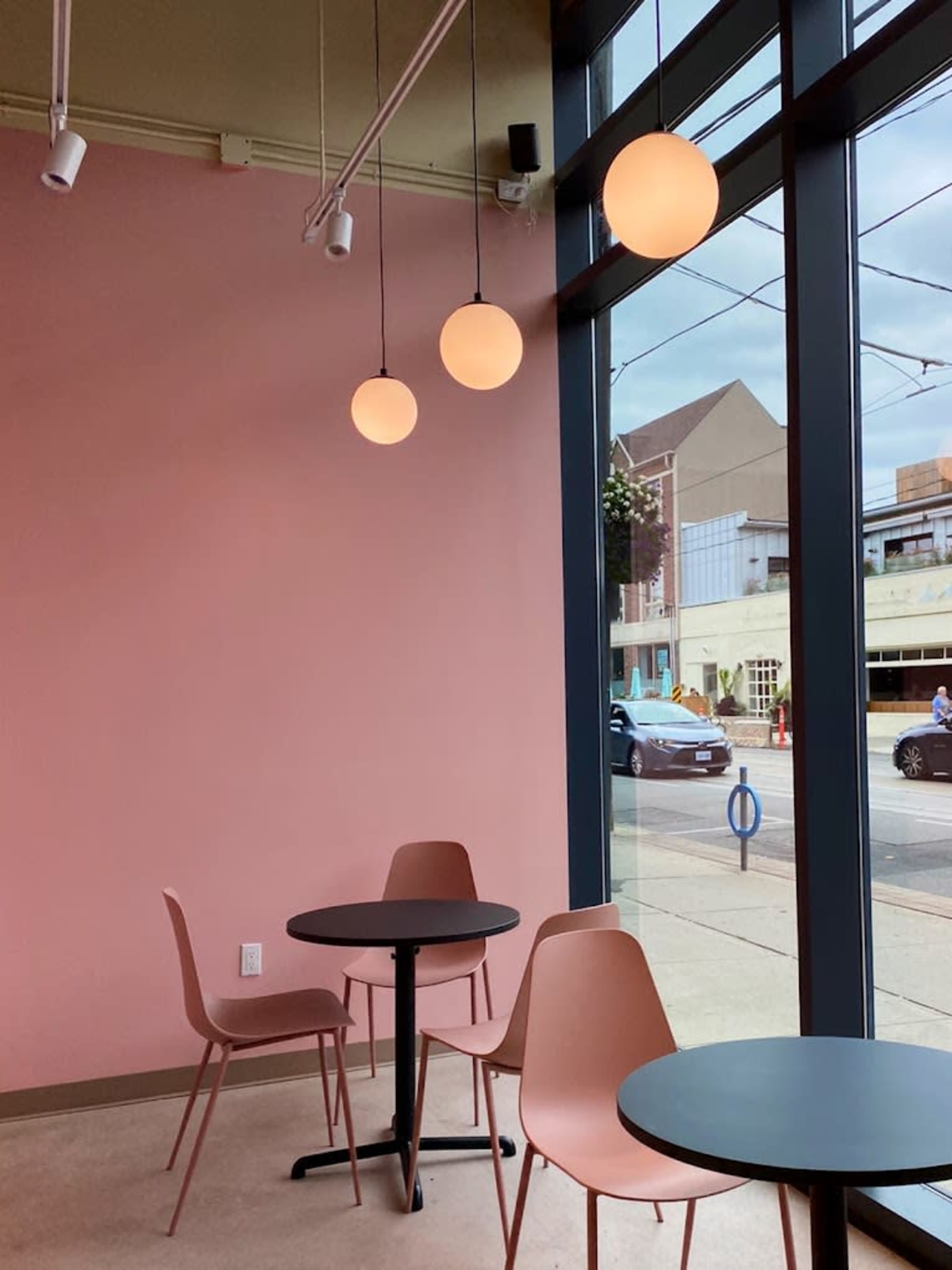 A café interior with three pendant lights hanging above a round table and two pink chairs, set against a pink wall and large windows facing the street.