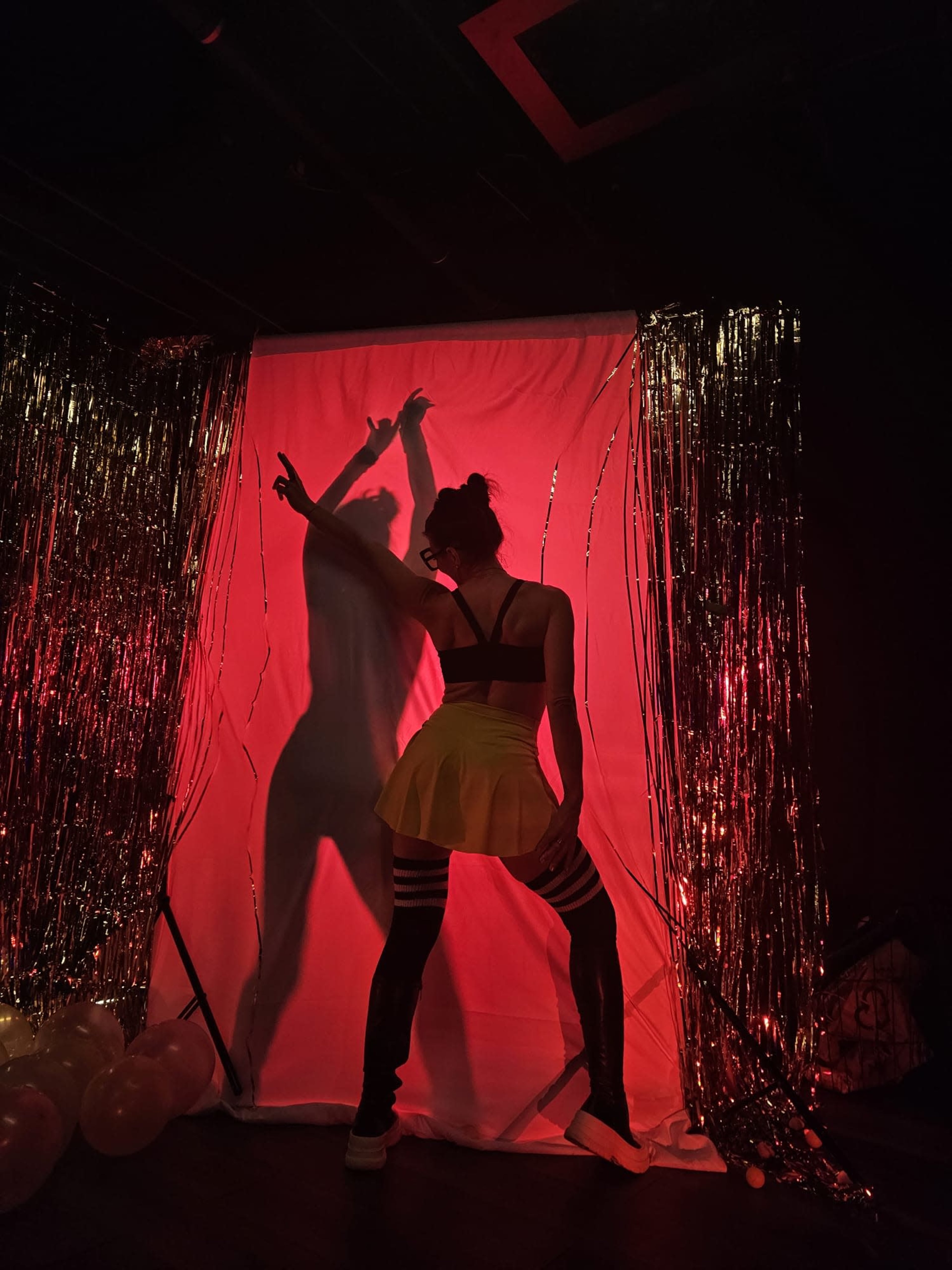 A silhouette of a person in a black top and a yellow skirt poses against a red-lit background with golden streamers.
