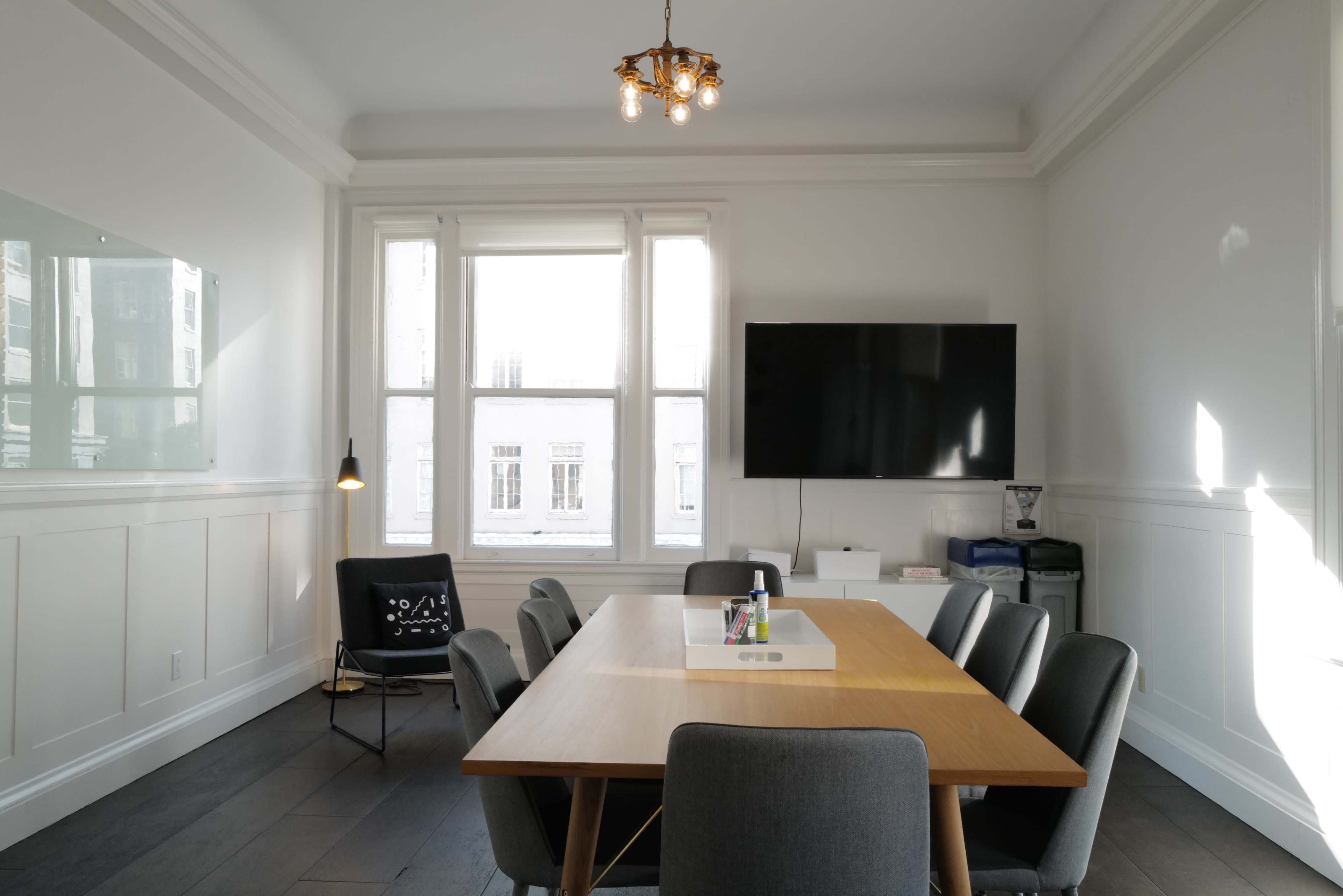 A modern conference room features a large wooden table surrounded by gray chairs, a wall-mounted television, and large windows providing ample natural light.