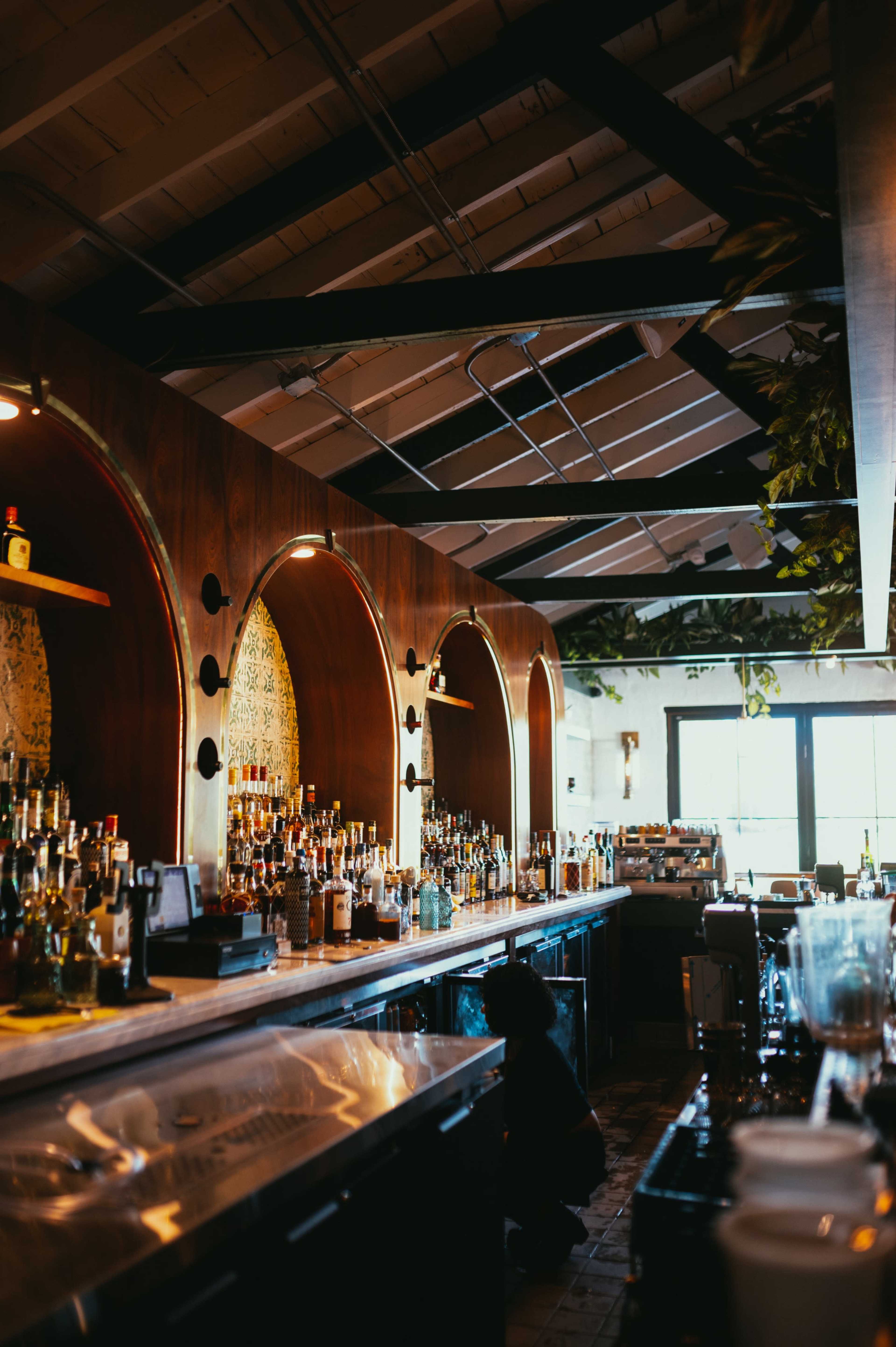 The image shows a bar area with a wooden counter, illuminated shelves displaying various bottles, and arched alcoves against a backdrop of greenery.