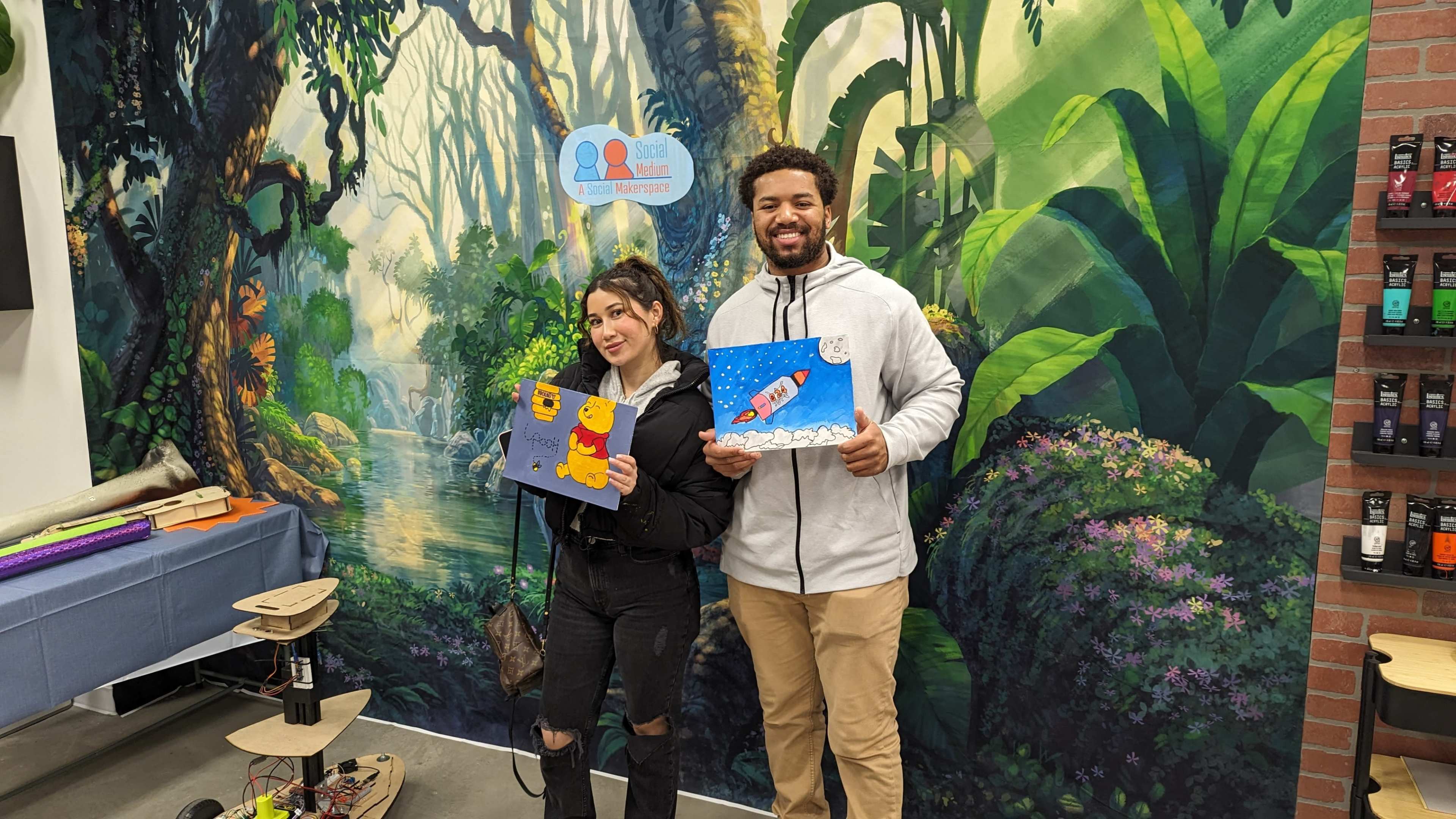 A man and a woman hold up their painted artwork in front of a vibrant mural depicting a lush, green forest.