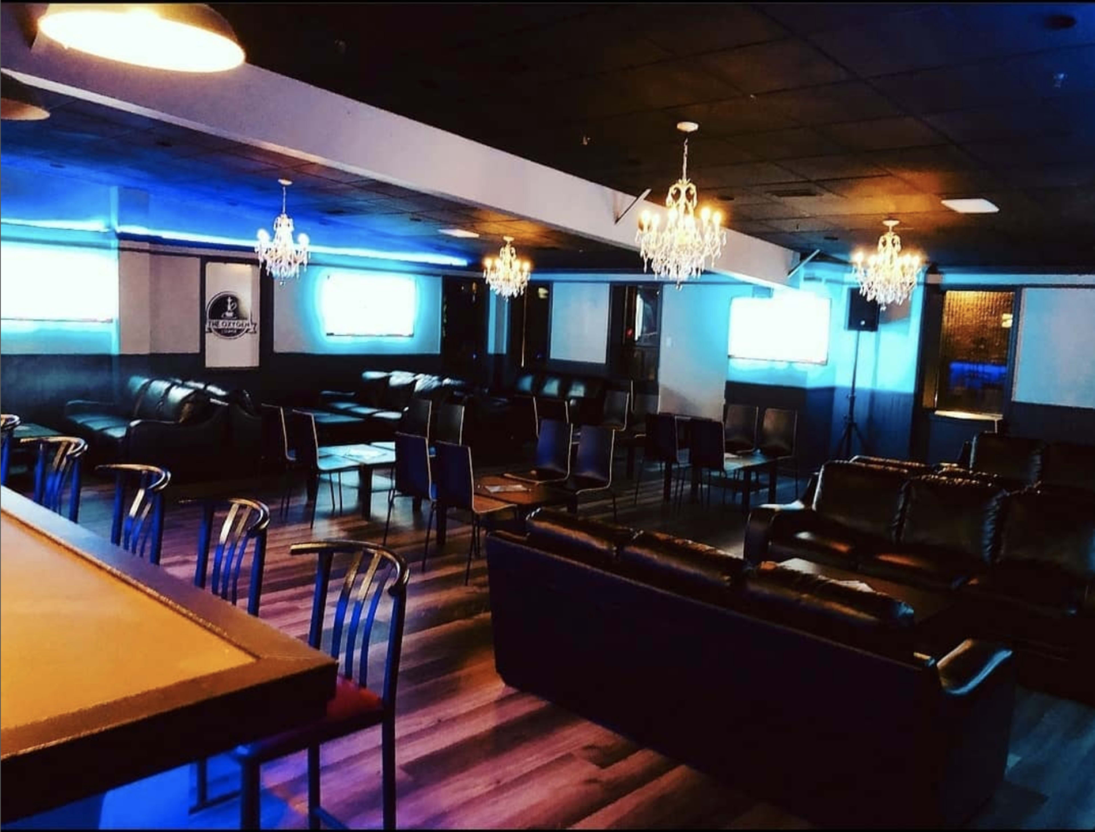 A dimly lit lounge area with leather seating, small tables, and chandeliers, featuring screens on the walls.