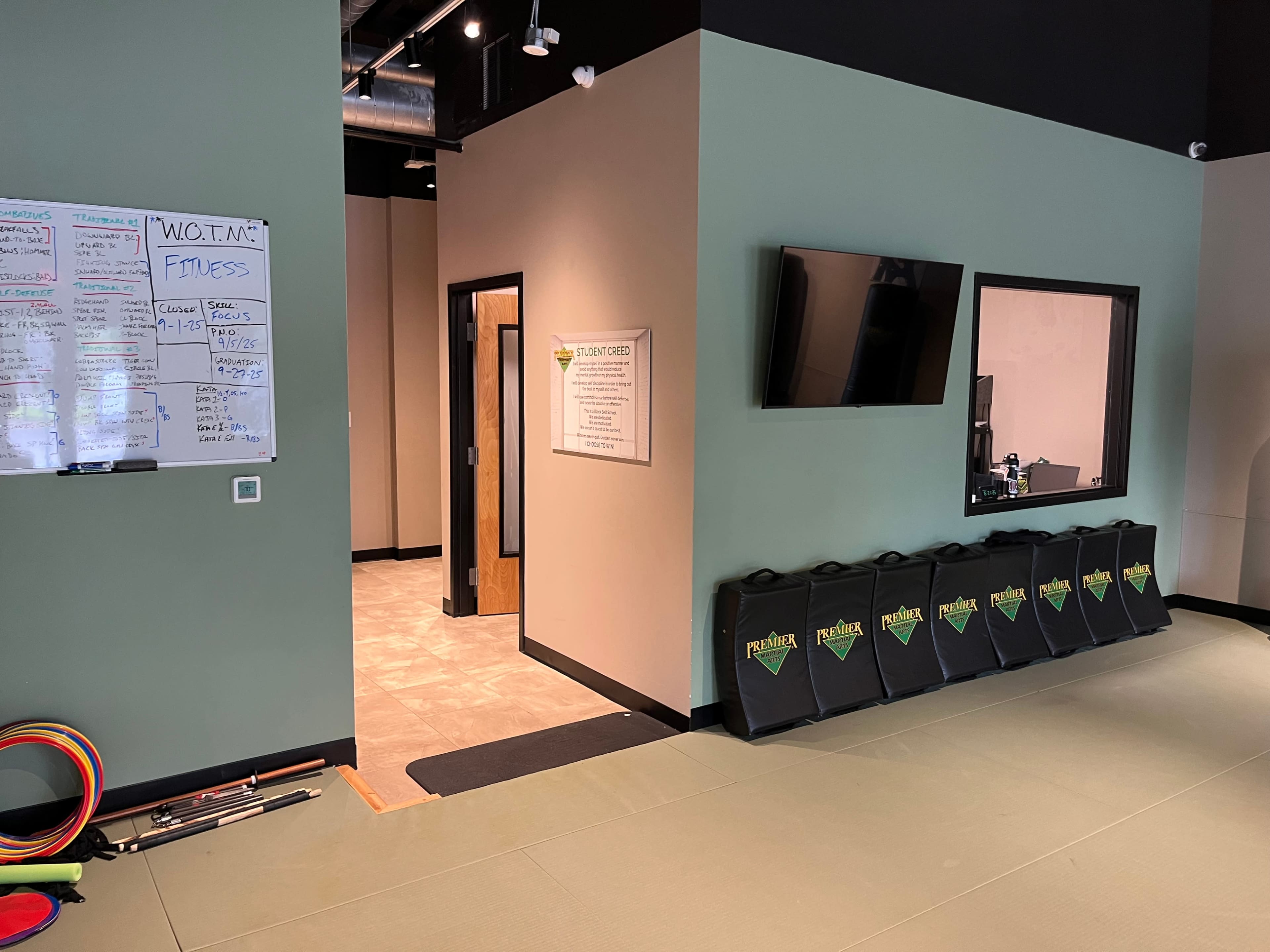 The image shows the interior of a fitness facility featuring a green and beige color scheme, a whiteboard with notes, a television on the wall, and a row of fitness mats lined up along the floor.