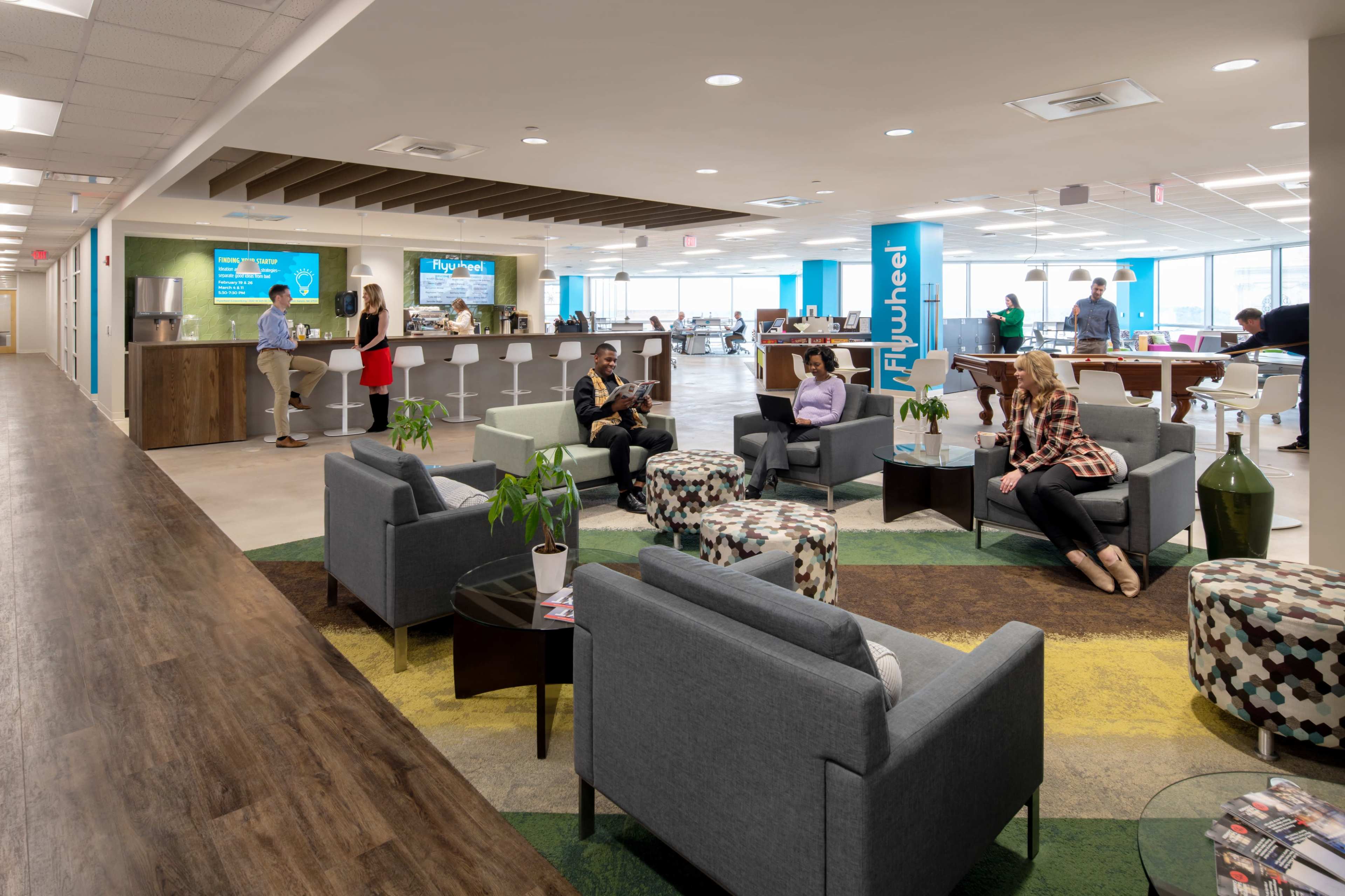 A modern office space with grouped seating areas, a communal kitchen, and workers engaging in various activities.