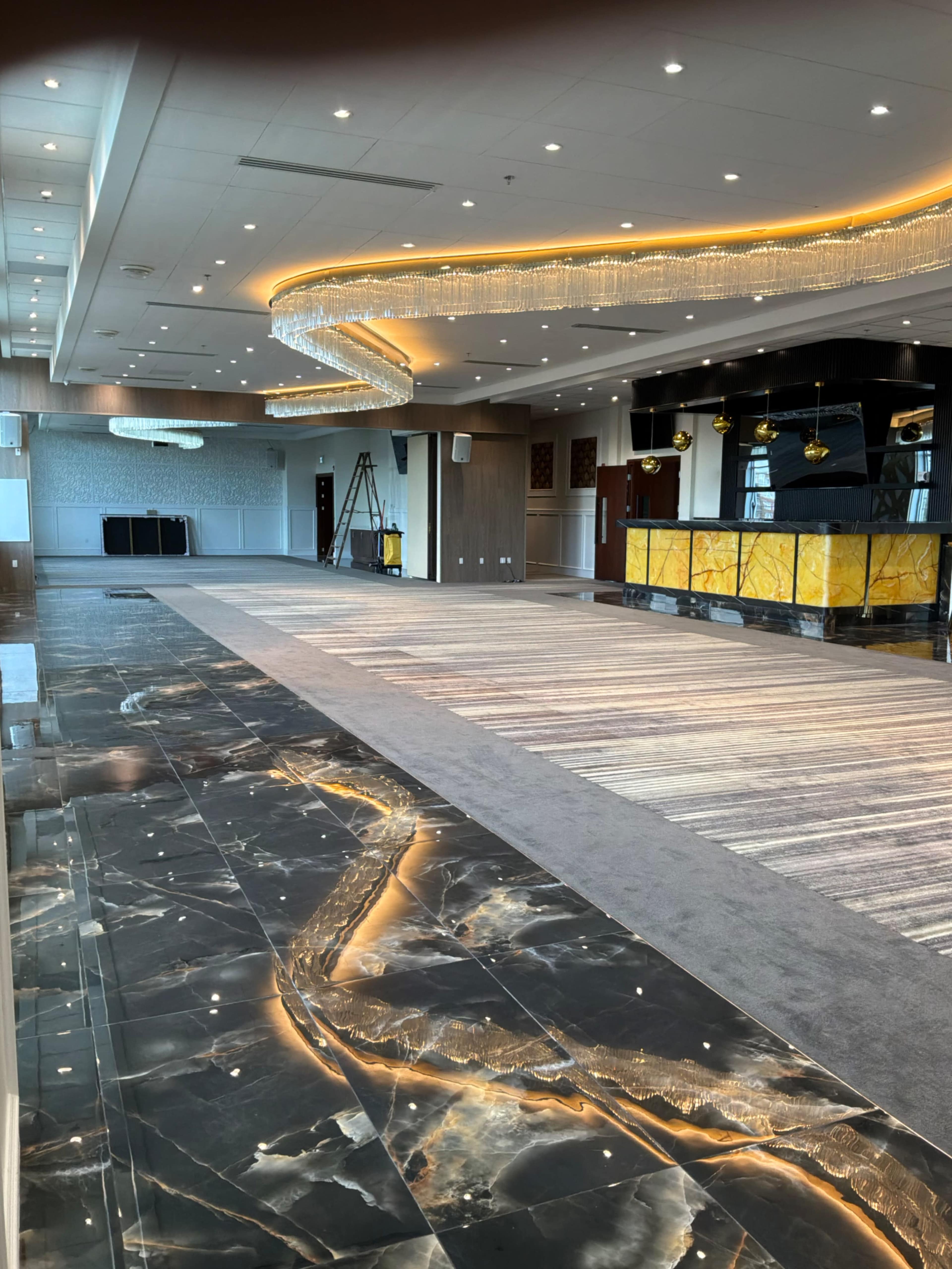 The image shows a spacious, elegantly designed interior of a banquet hall featuring polished marble floors, decorative lighting fixtures, and a reception area with a counter.