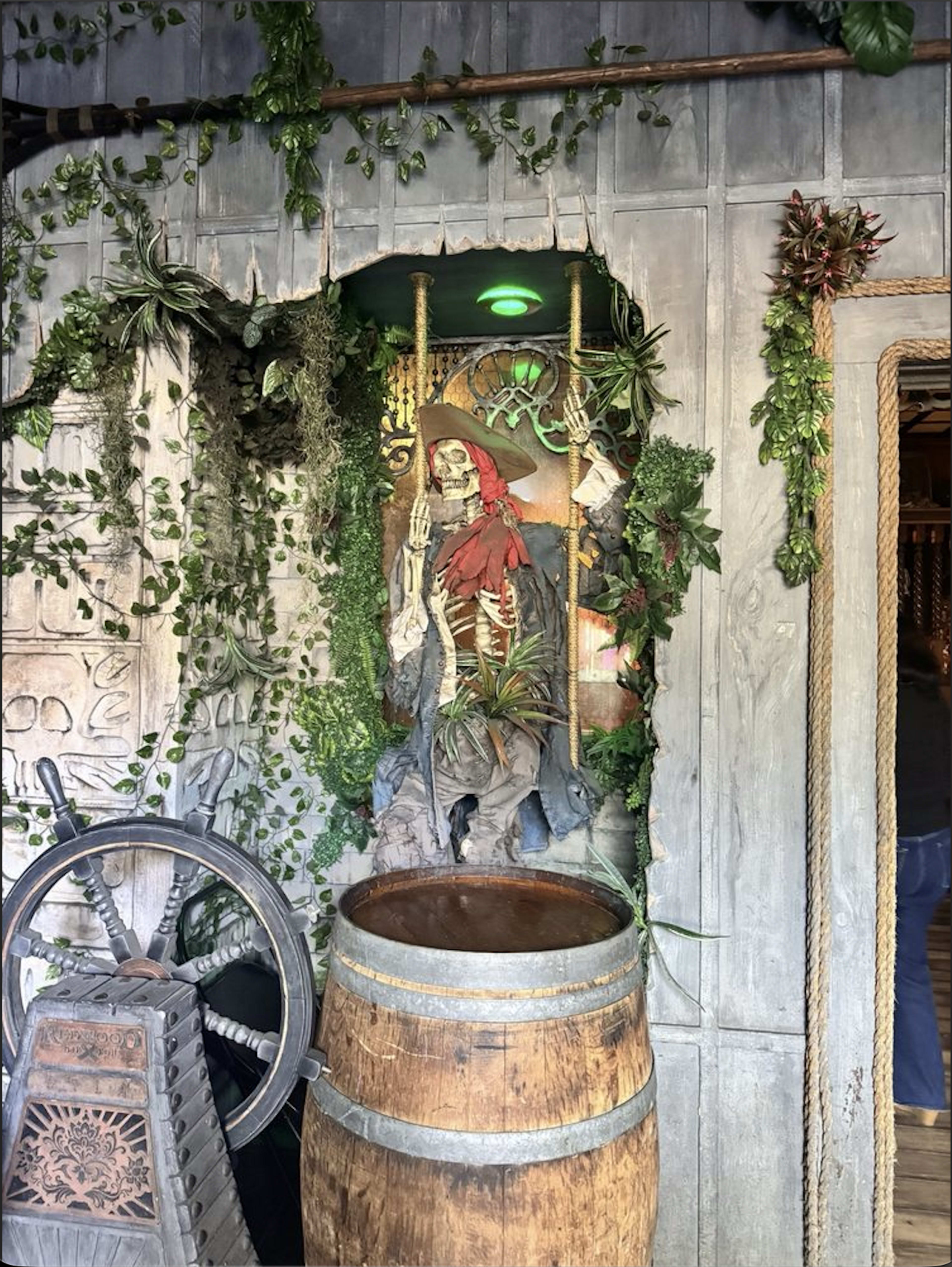 The image shows a wooden barrel next to a decorative wall featuring a skeleton surrounded by plants and greenery, illuminated by a green light.