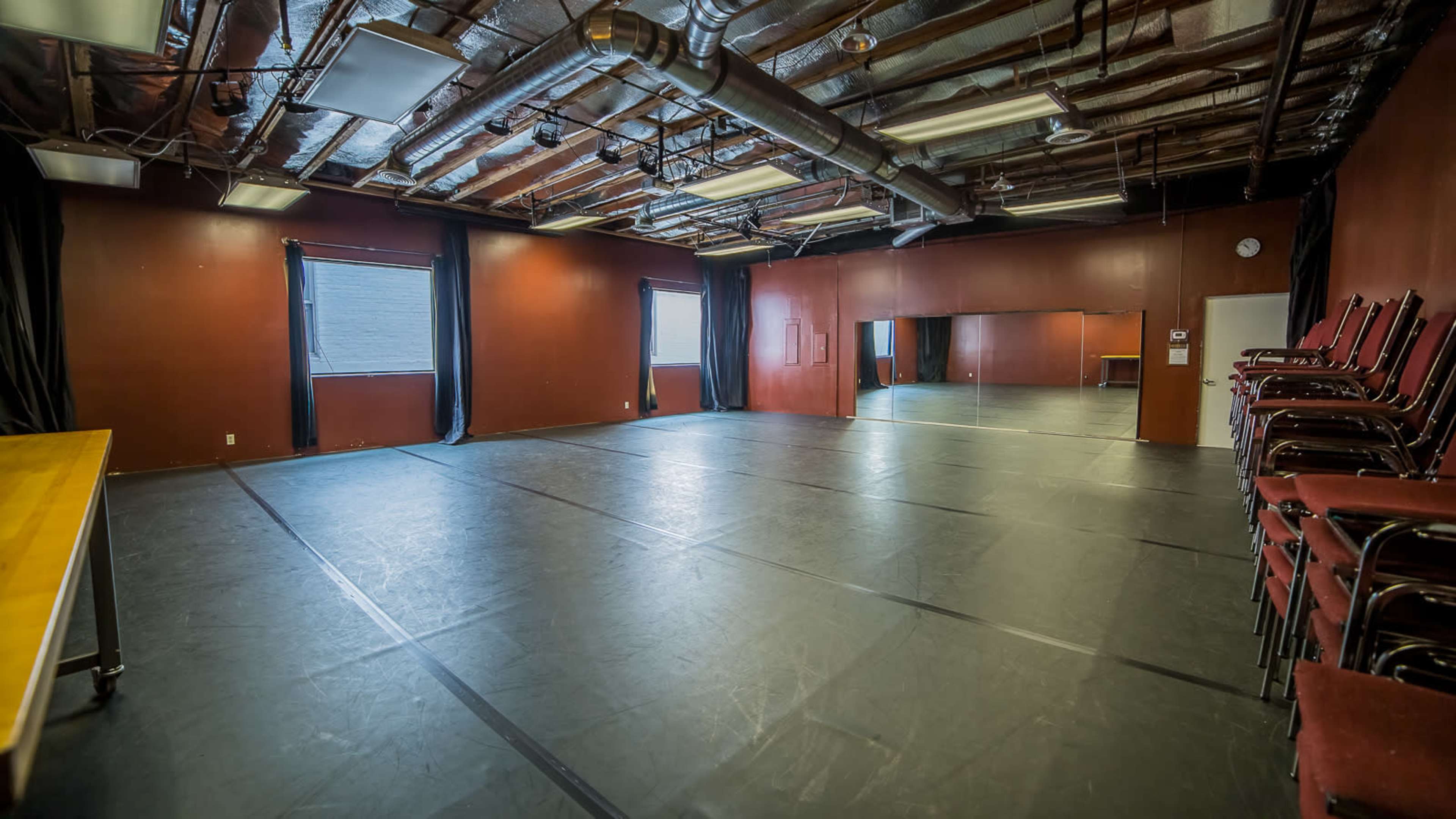 The image shows an empty studio space with dark walls, large windows, and a mirrored wall, featuring a stack of red chairs on one side.