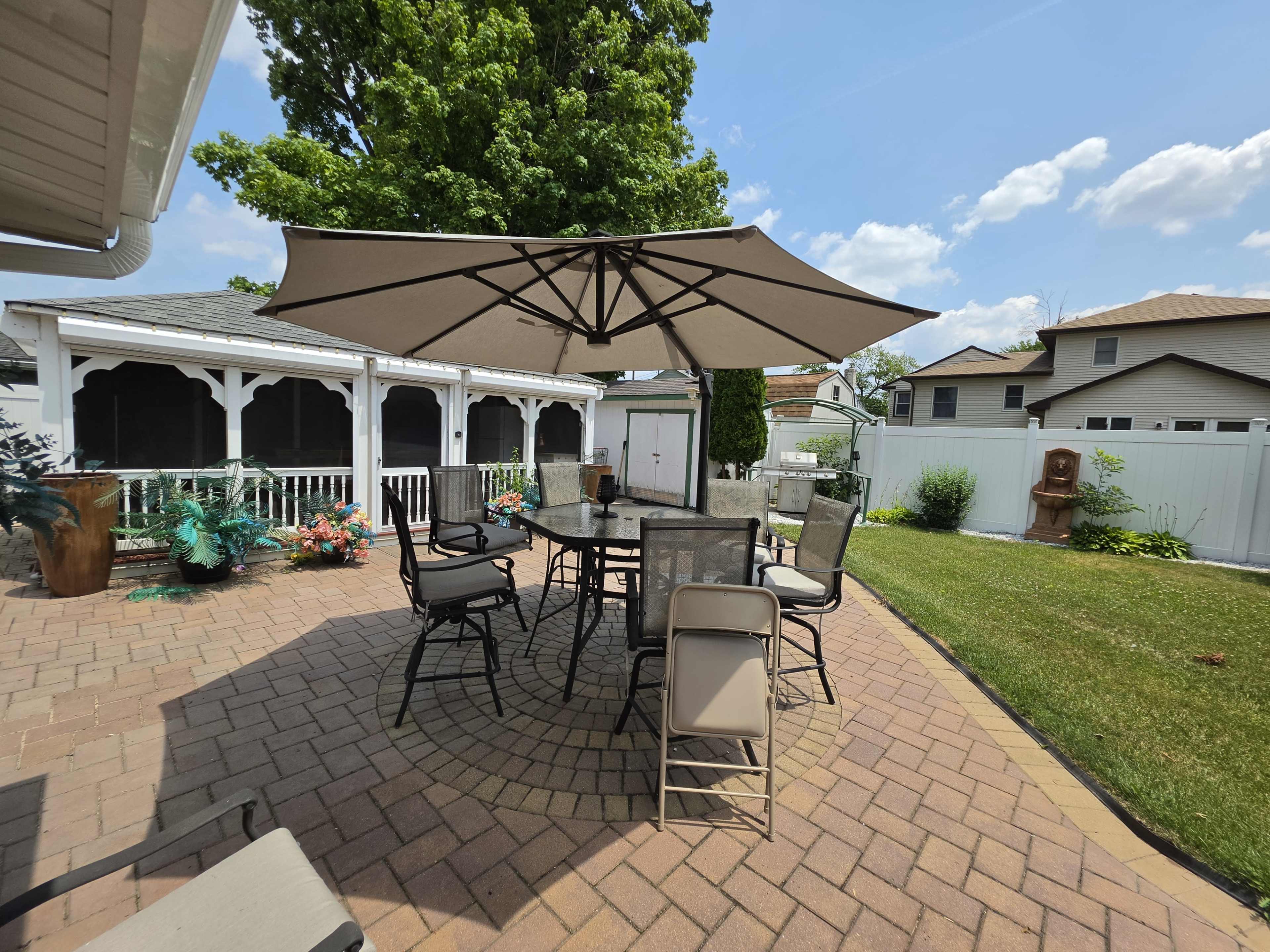 The image shows a patio area with a large umbrella over a dining table surrounded by chairs, set against a backdrop of a white fence and a garden.