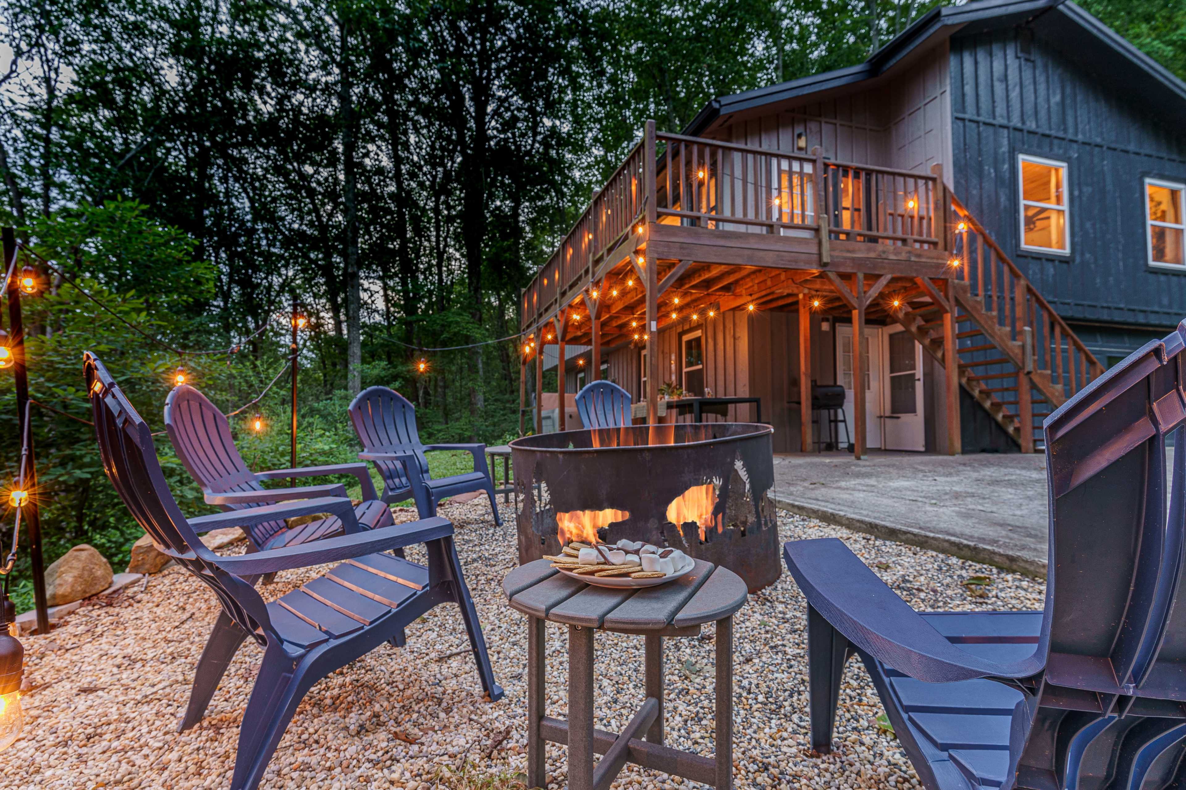 A stone patio with a fire pit and several blue Adirondack chairs is surrounded by trees, while a two-story wooden cabin with string lights illuminates the scene in the background.