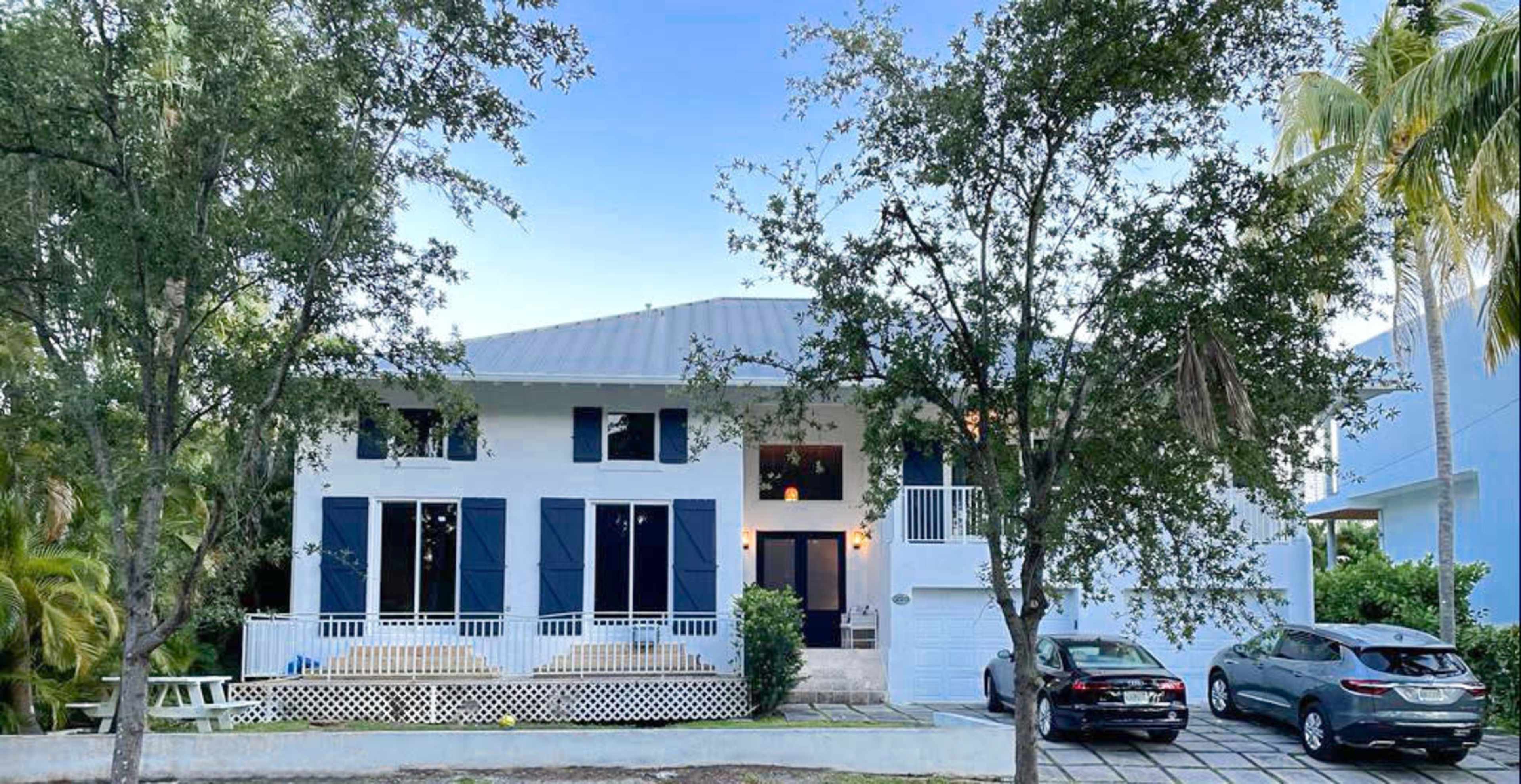 A two-story modern house with a gray metal roof and blue shutters is flanked by palm trees, showcasing a driveway with two parked cars.