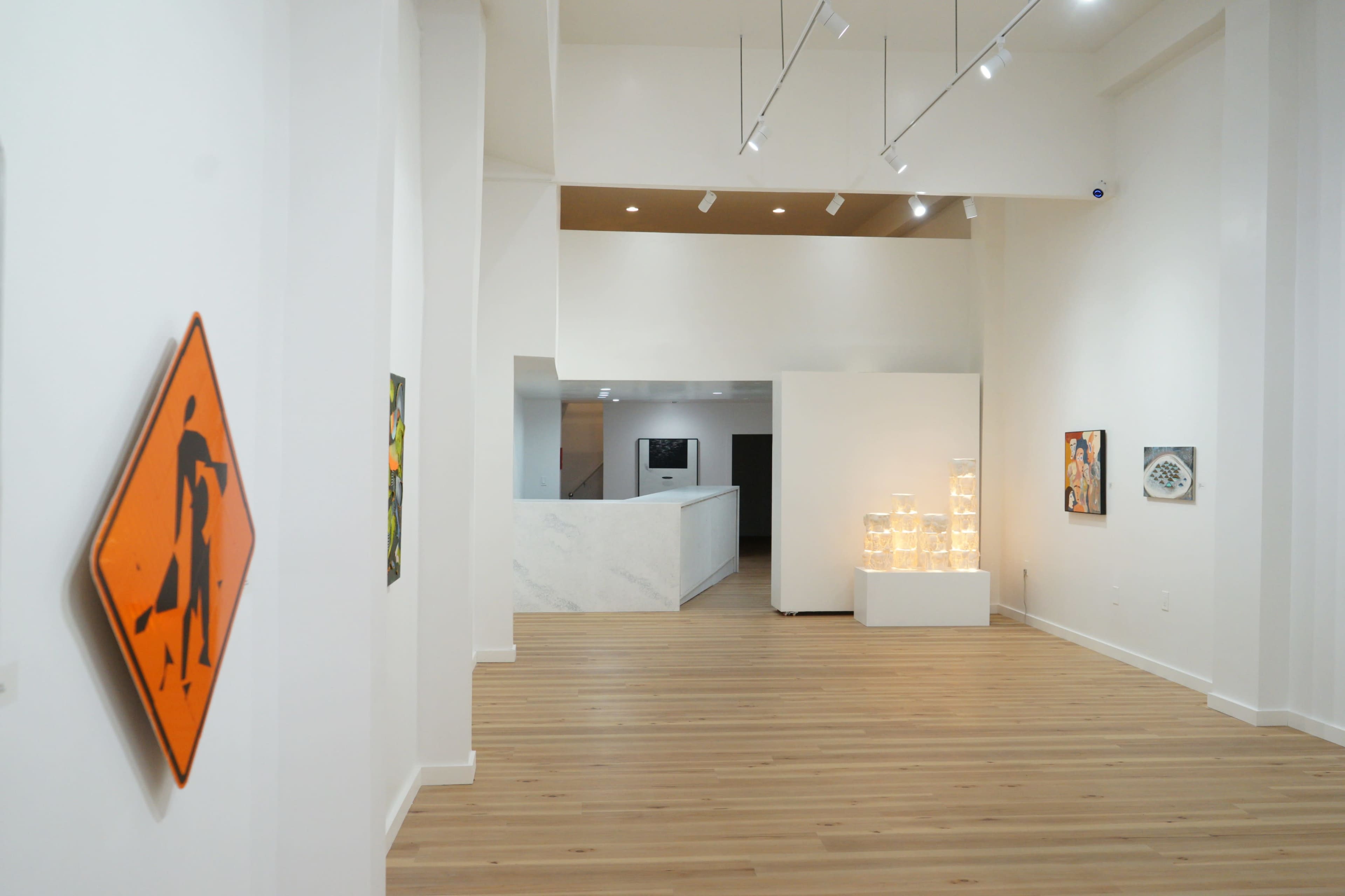 The image shows the interior of a contemporary art gallery featuring wooden flooring, white walls, and a selection of displayed artworks, including an orange construction sign.