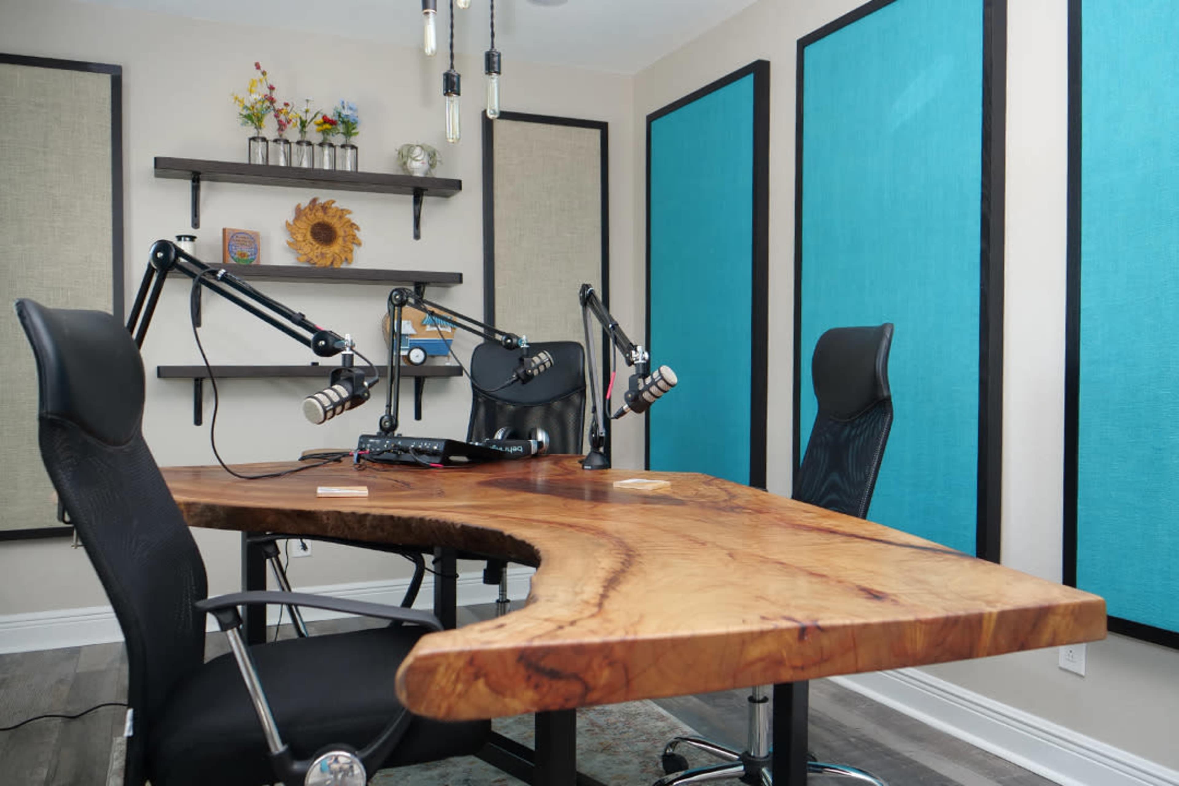 The image shows a modern podcast recording studio featuring a large wooden table with two black office chairs and adjustable microphone arms, surrounded by blue soundproofing panels on the walls.