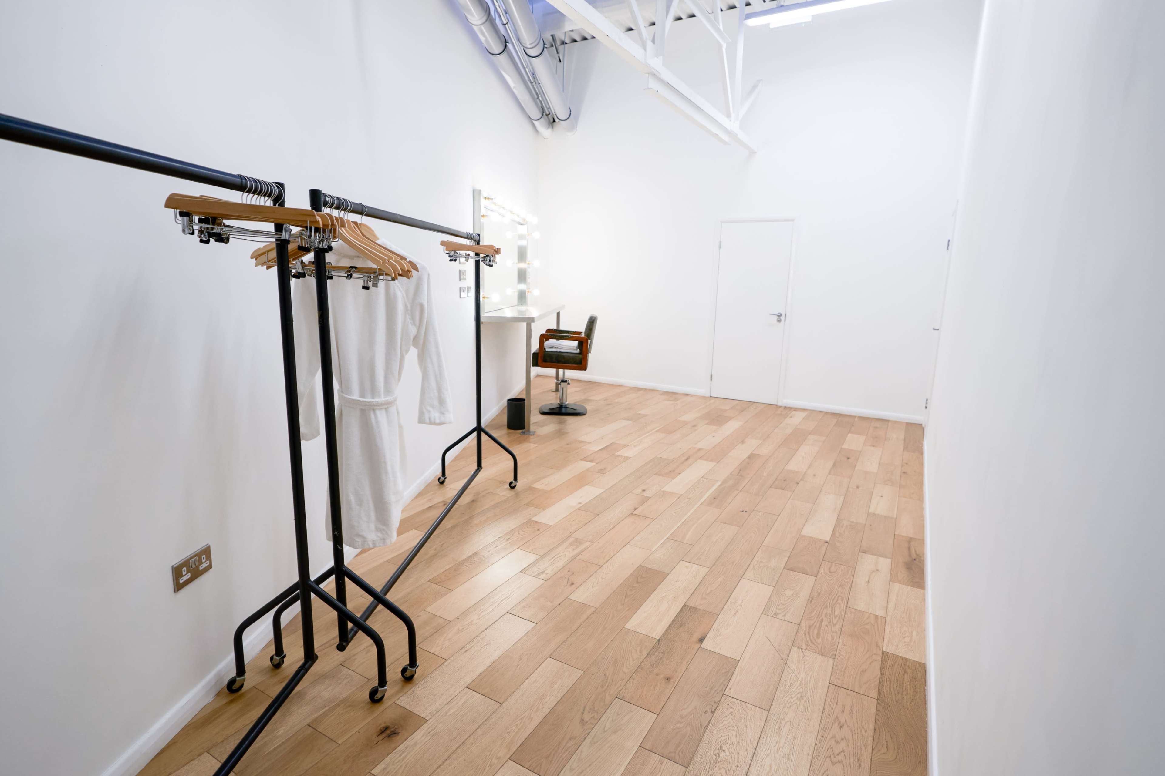 The image shows an empty room with wooden flooring, a clothing rack with hangers, a styling chair, and a mirror against a white wall.