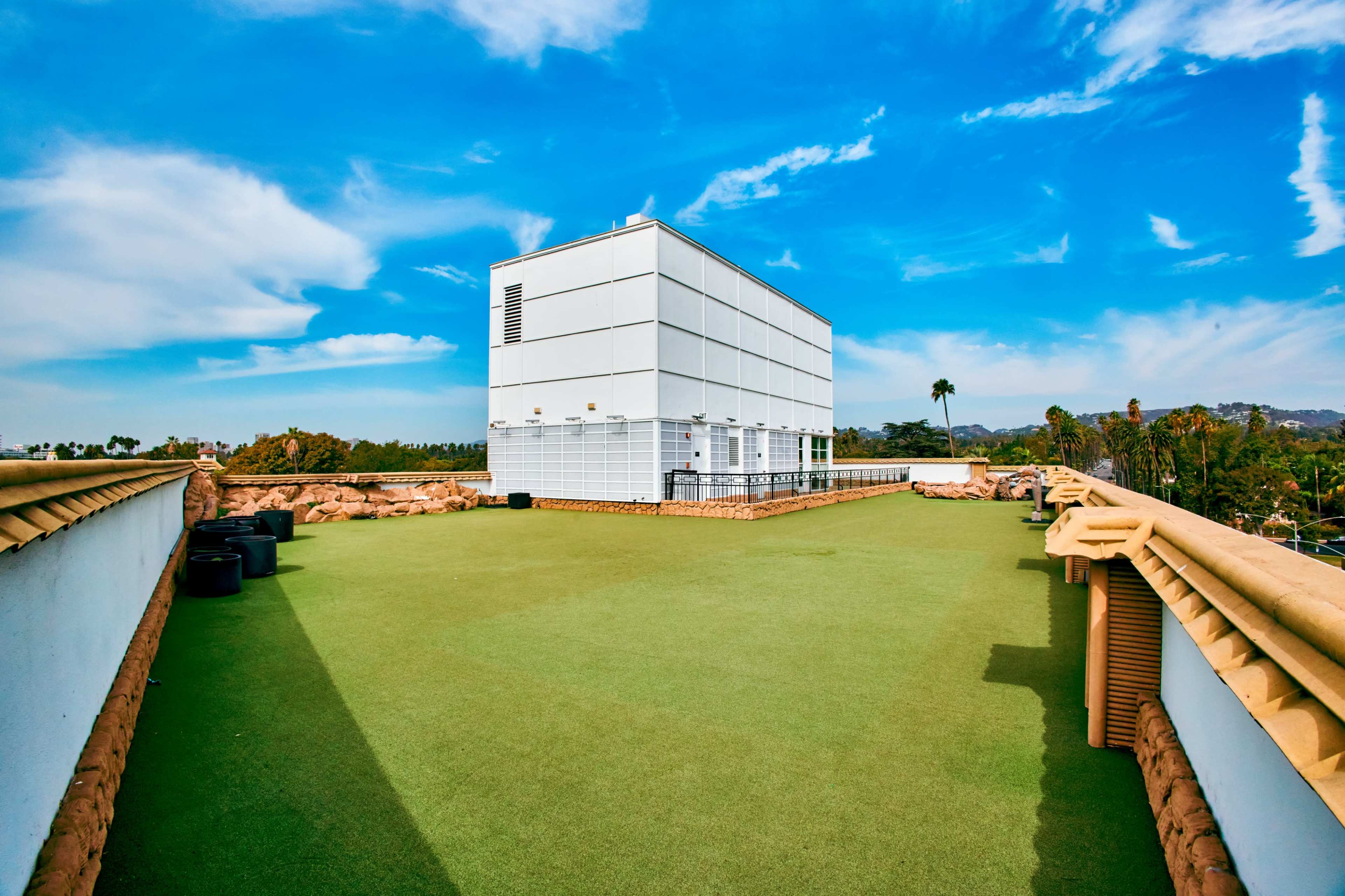A large, white building with a flat roof sits atop a green, artificial turf area against a backdrop of blue sky and wispy clouds.