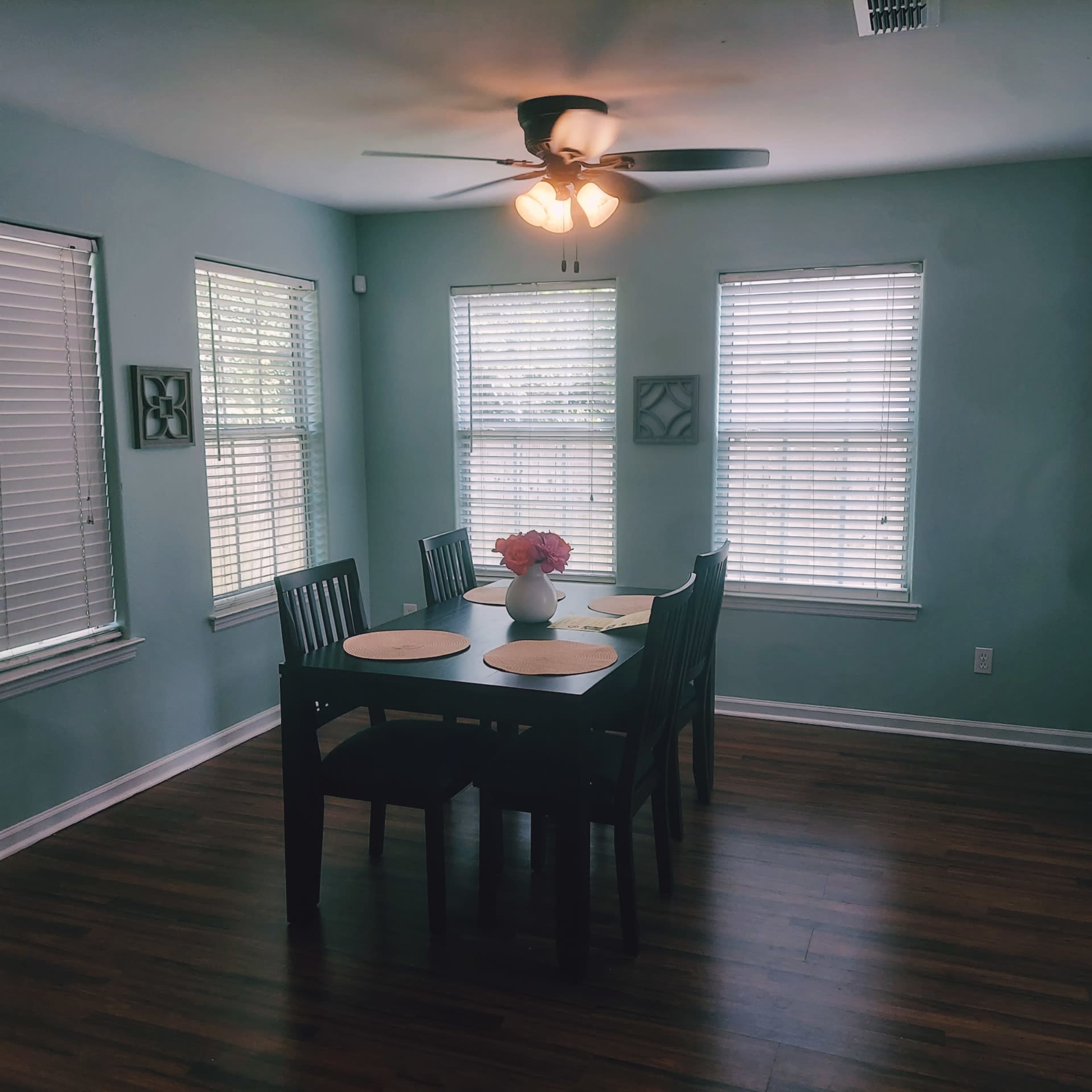 A dining area with a black table and chairs is set near windows with blinds, featuring a vase of flowers on the table and a ceiling fan overhead.
