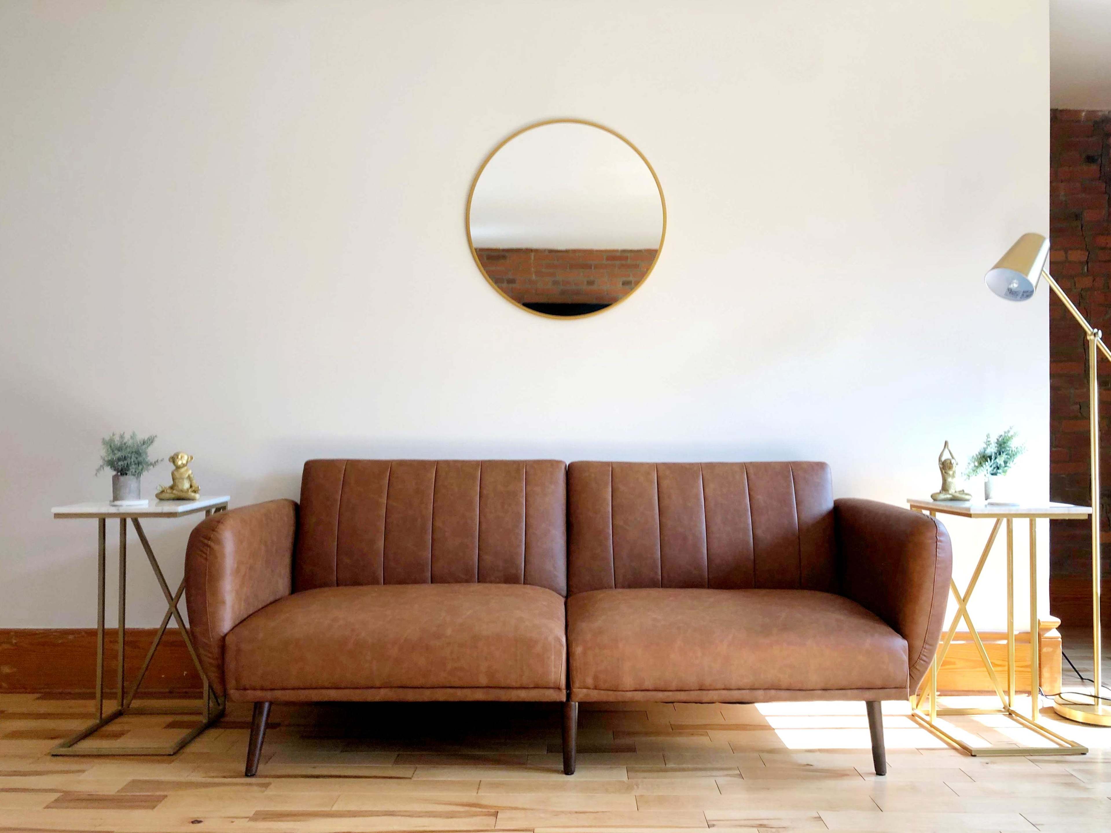 A brown leather sofa is placed between two gold side tables with small plants, and a round mirror hangs on the wall above it.