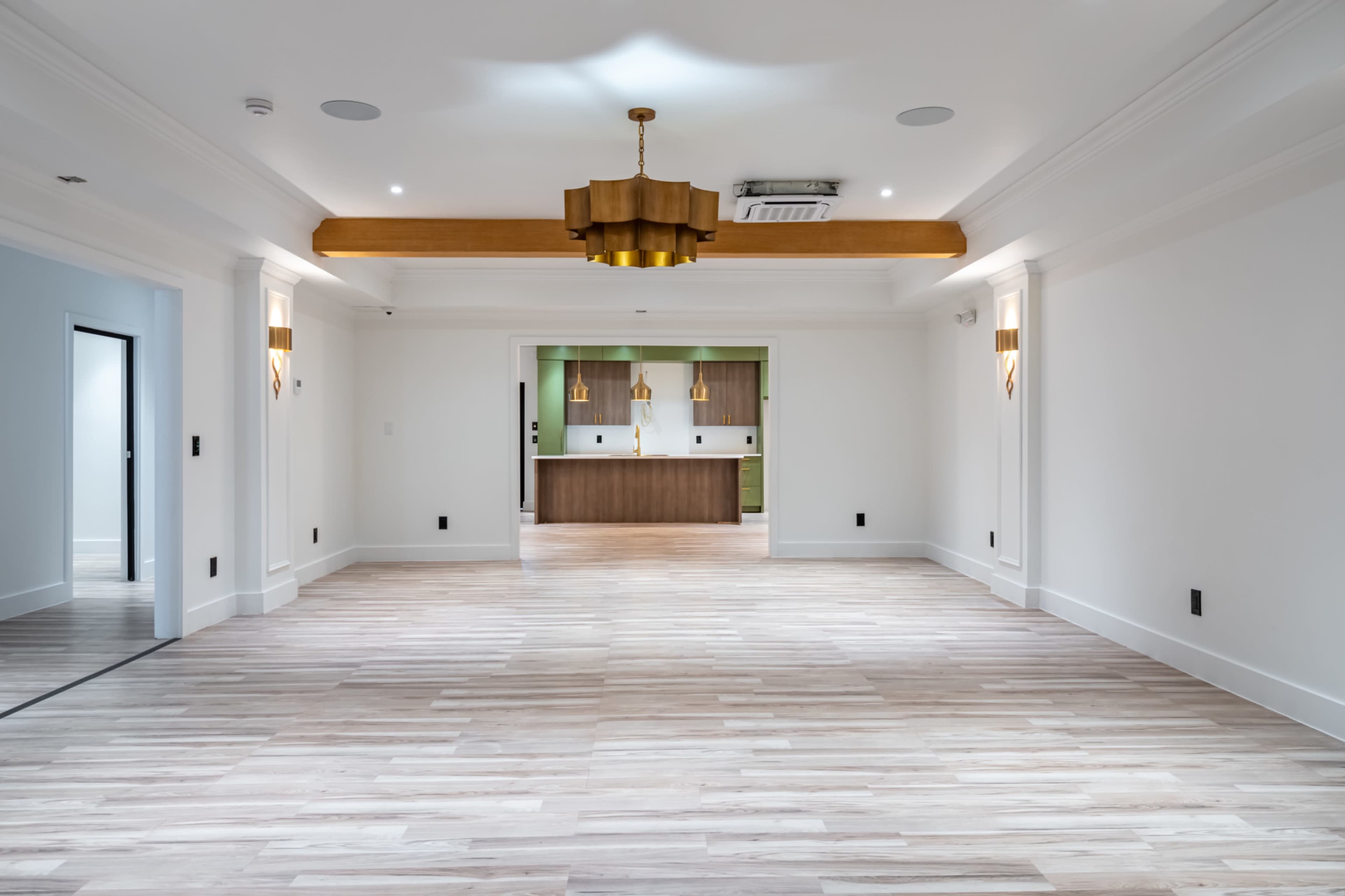 The image shows a spacious, well-lit room with light wooden flooring and a modern chandelier, leading into a kitchen area with wooden cabinetry visible in the background.