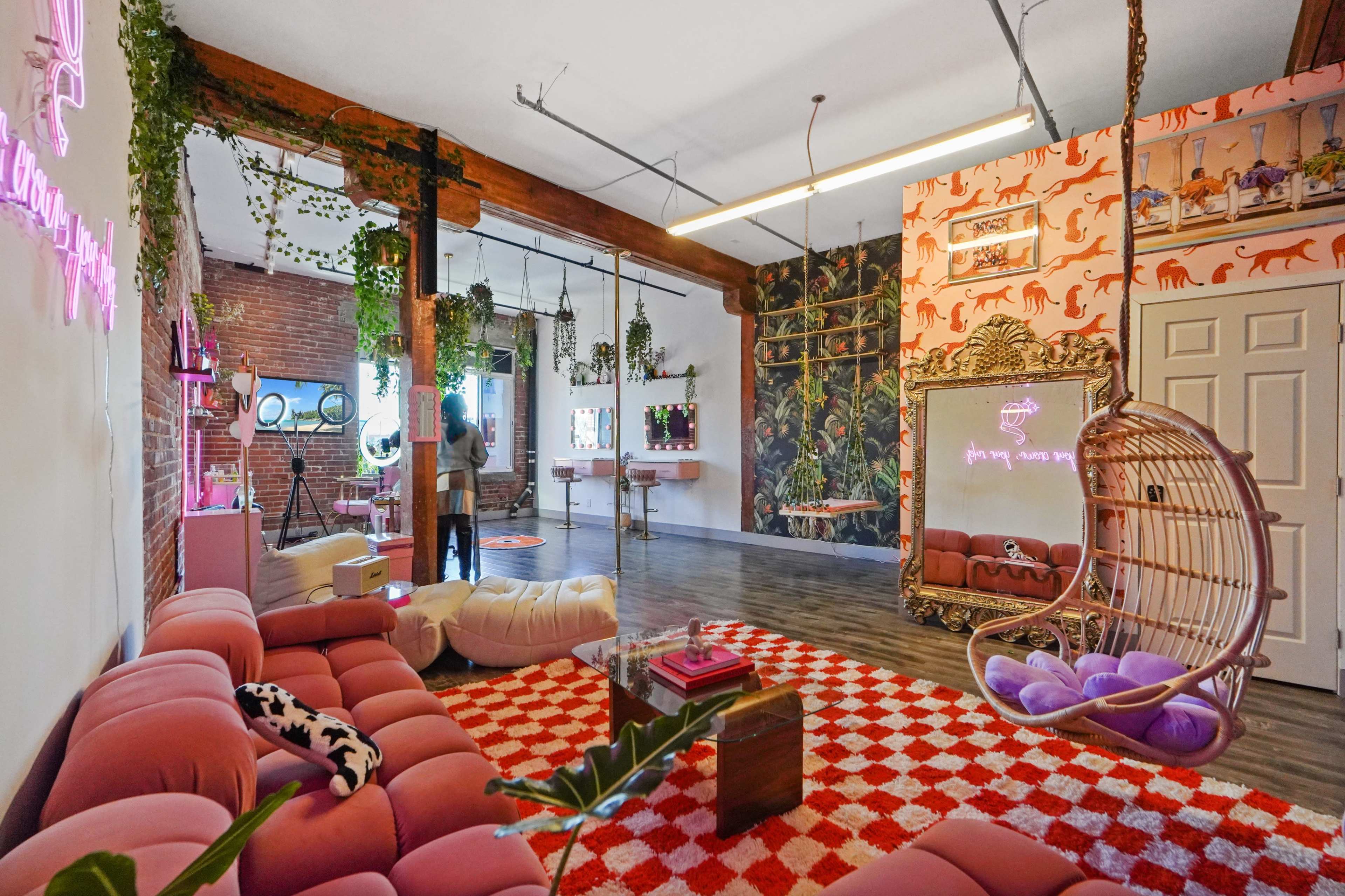 A colorful room featuring a pink sofa, a patterned red and white rug, a swinging chair, and various plants along the walls.