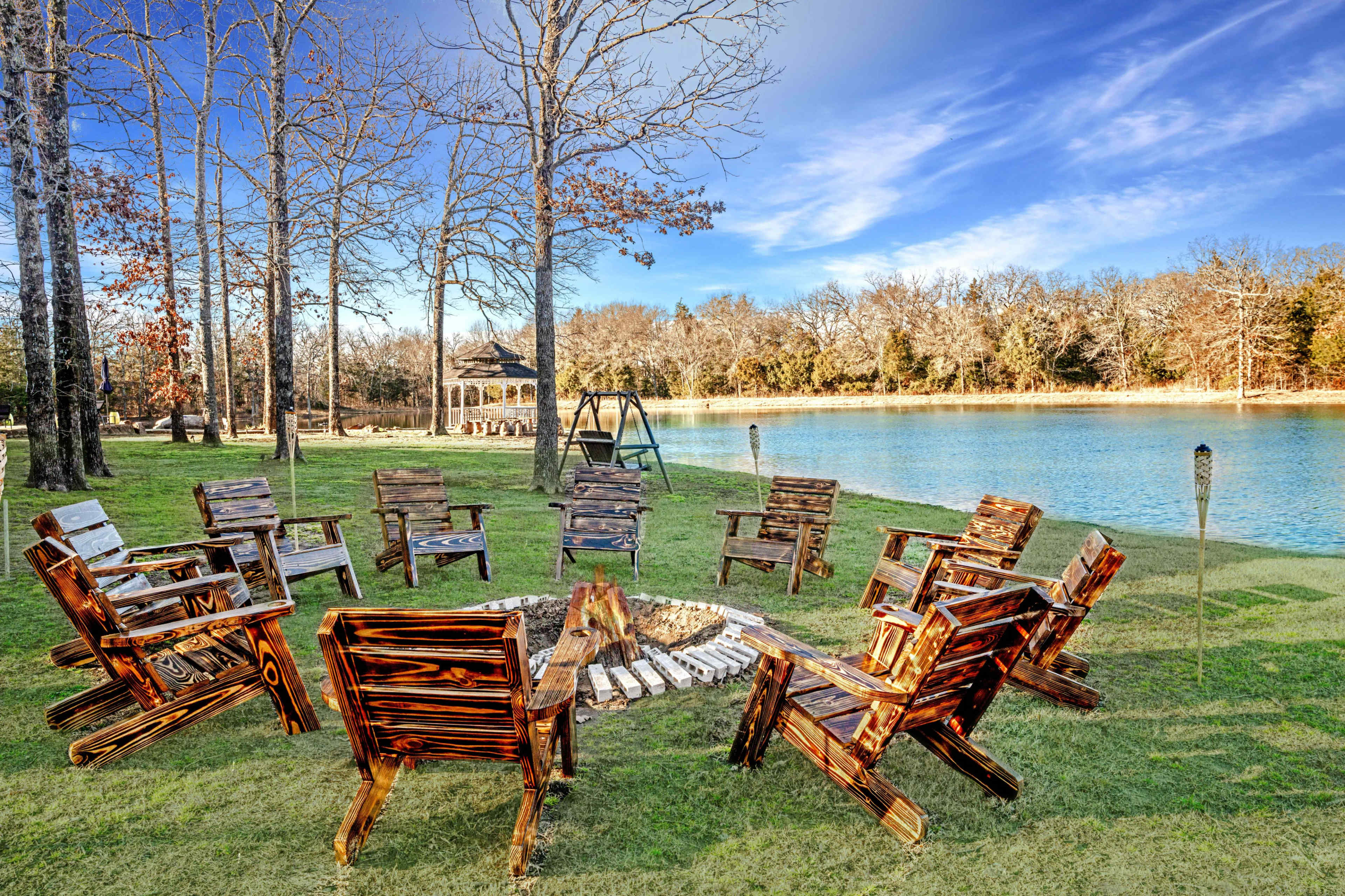 A circle of wooden chairs surrounds a fire pit near a calm lake, with trees and clear blue skies in the background.