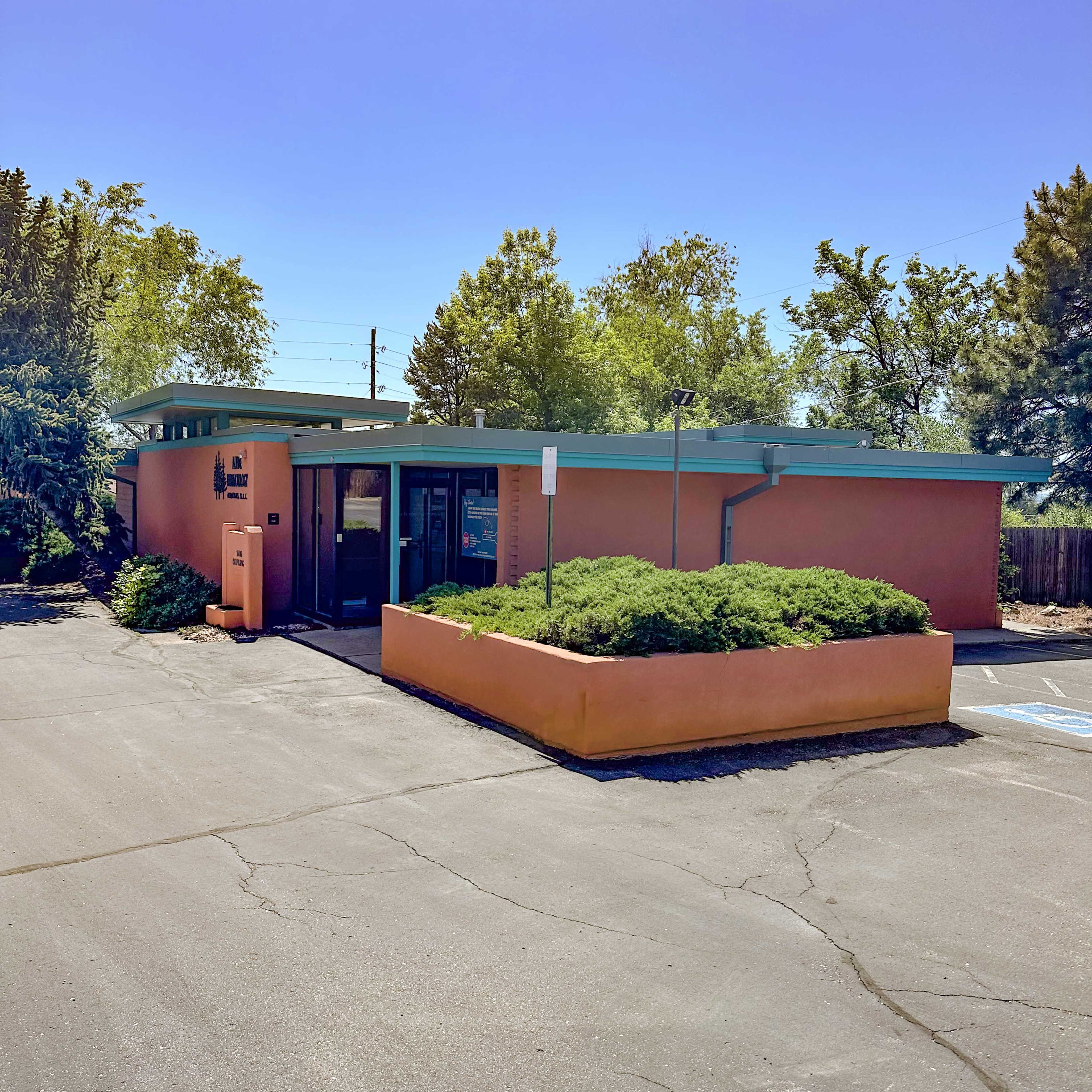 The image depicts a single-story, pink building with a turquoise roof, surrounded by green shrubbery and located in a parking area.