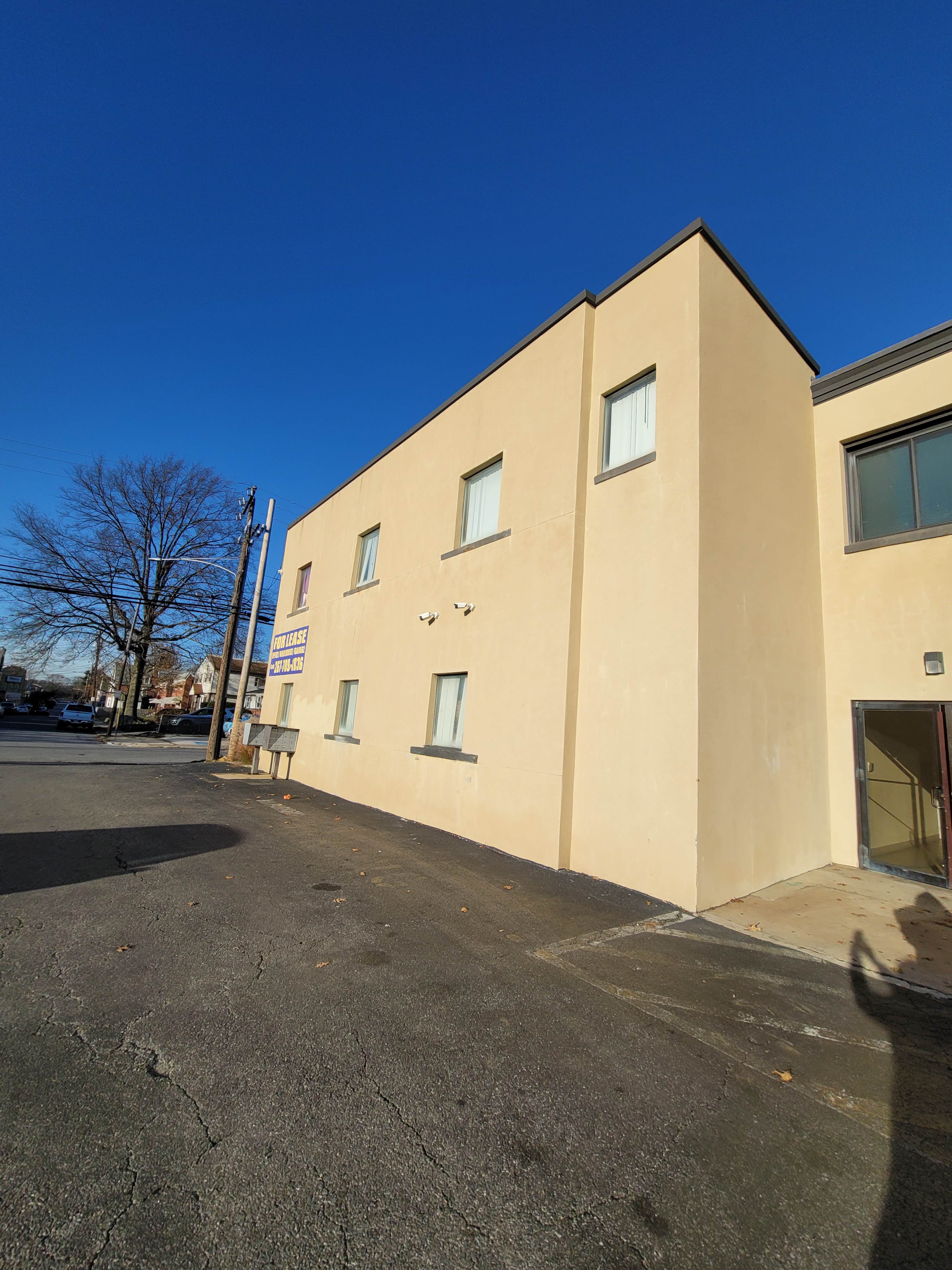 A beige building with multiple windows is shown under a clear blue sky, alongside an asphalt parking area.