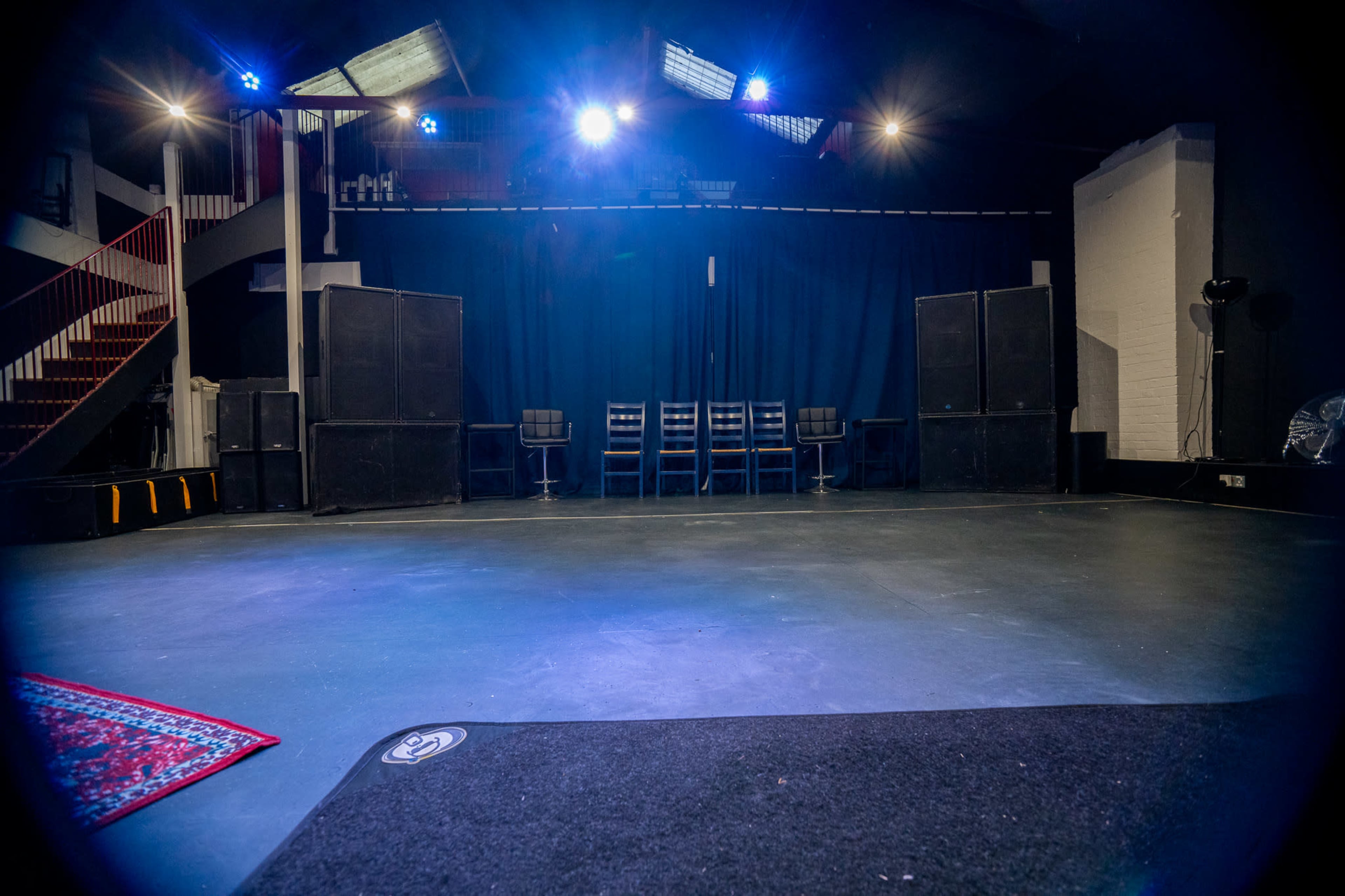 The image shows a stage area with a dark floor, sound speakers on either side, and several chairs arranged in a row against a curtain backdrop.