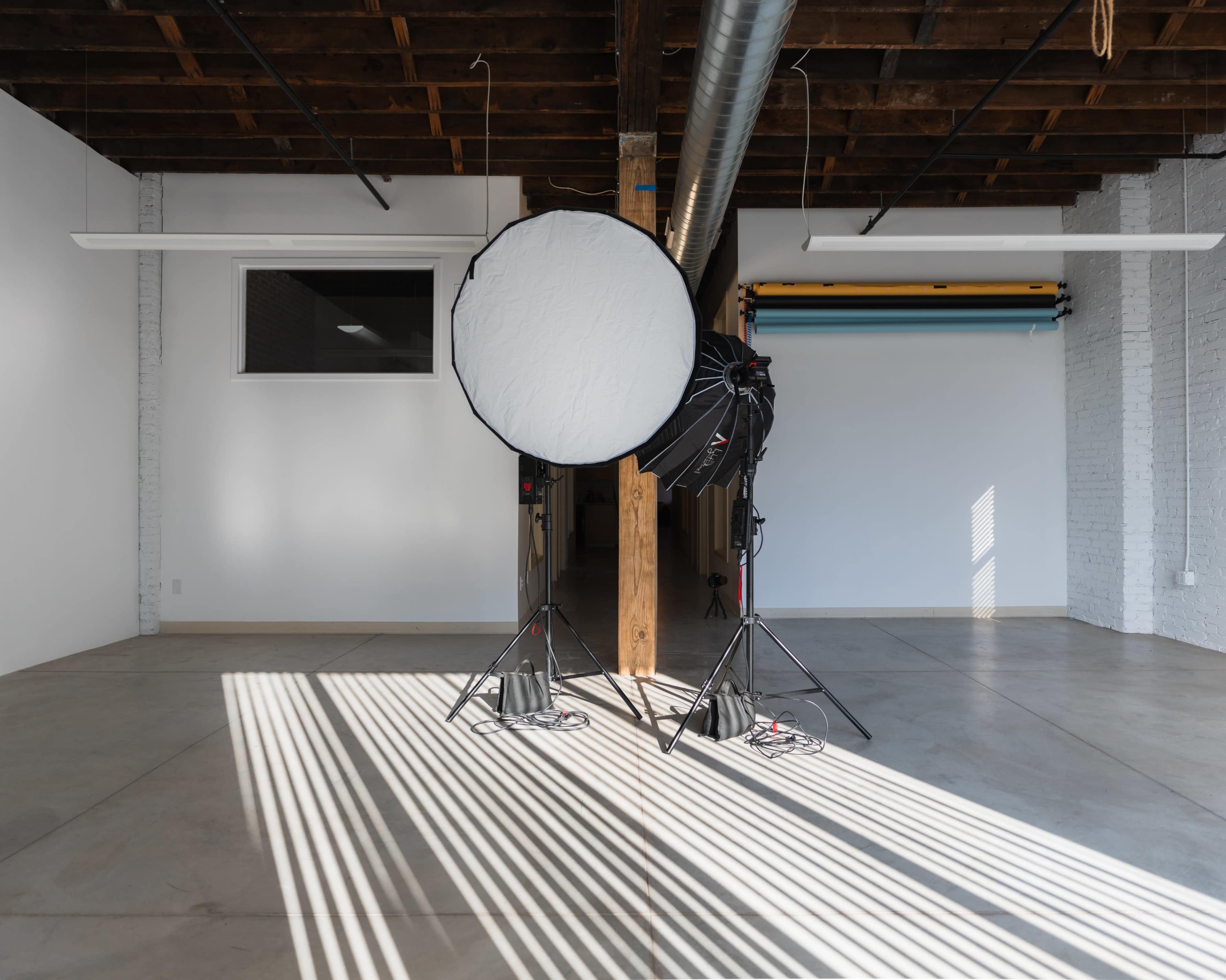 A photography studio features two light stands with diffusers set up on a concrete floor, casting shadows from overhead beams.