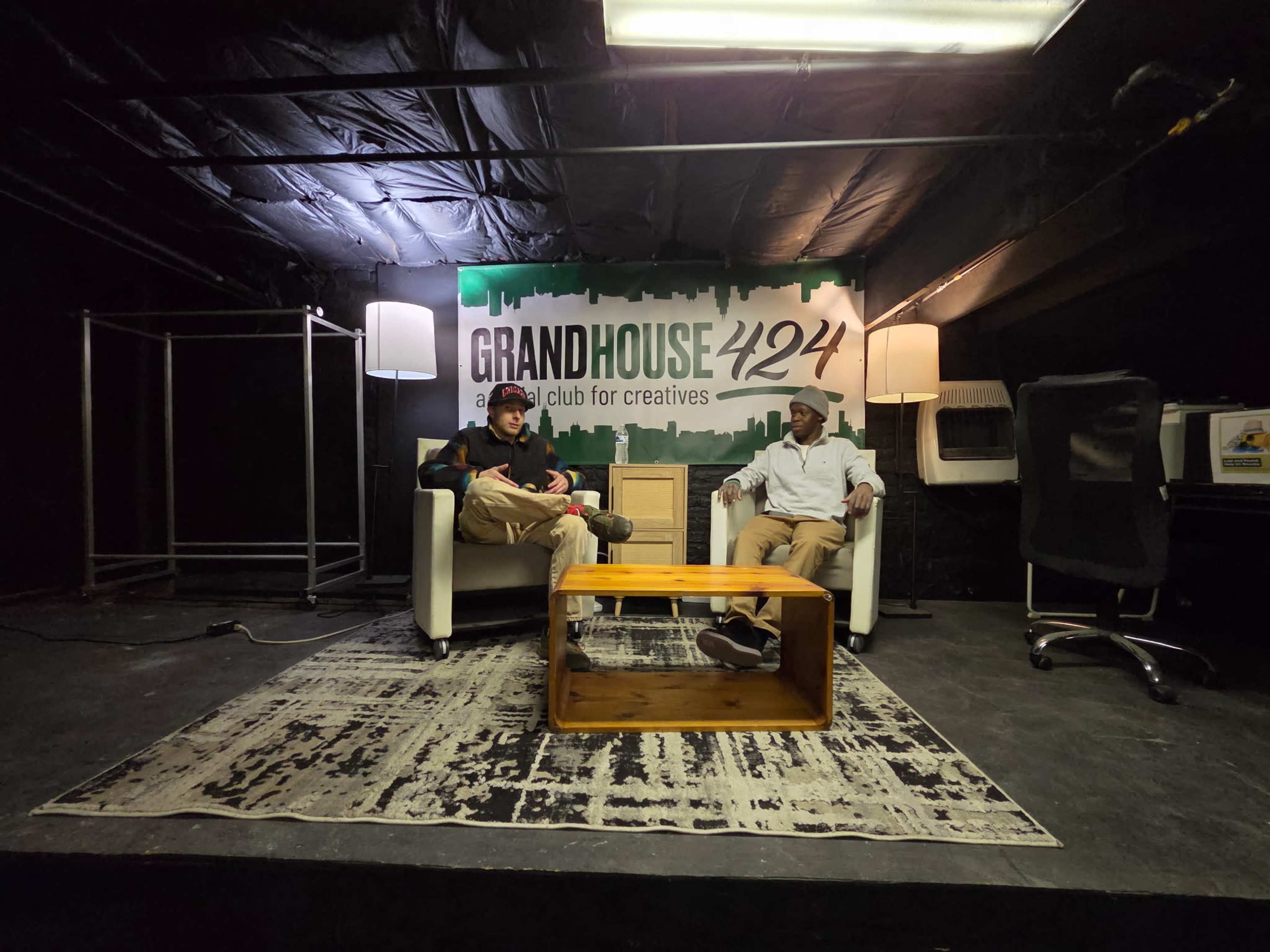 Two people are seated on white armchairs in a dimly lit studio space with a "GRANDHOUSE 424" sign on the wall.