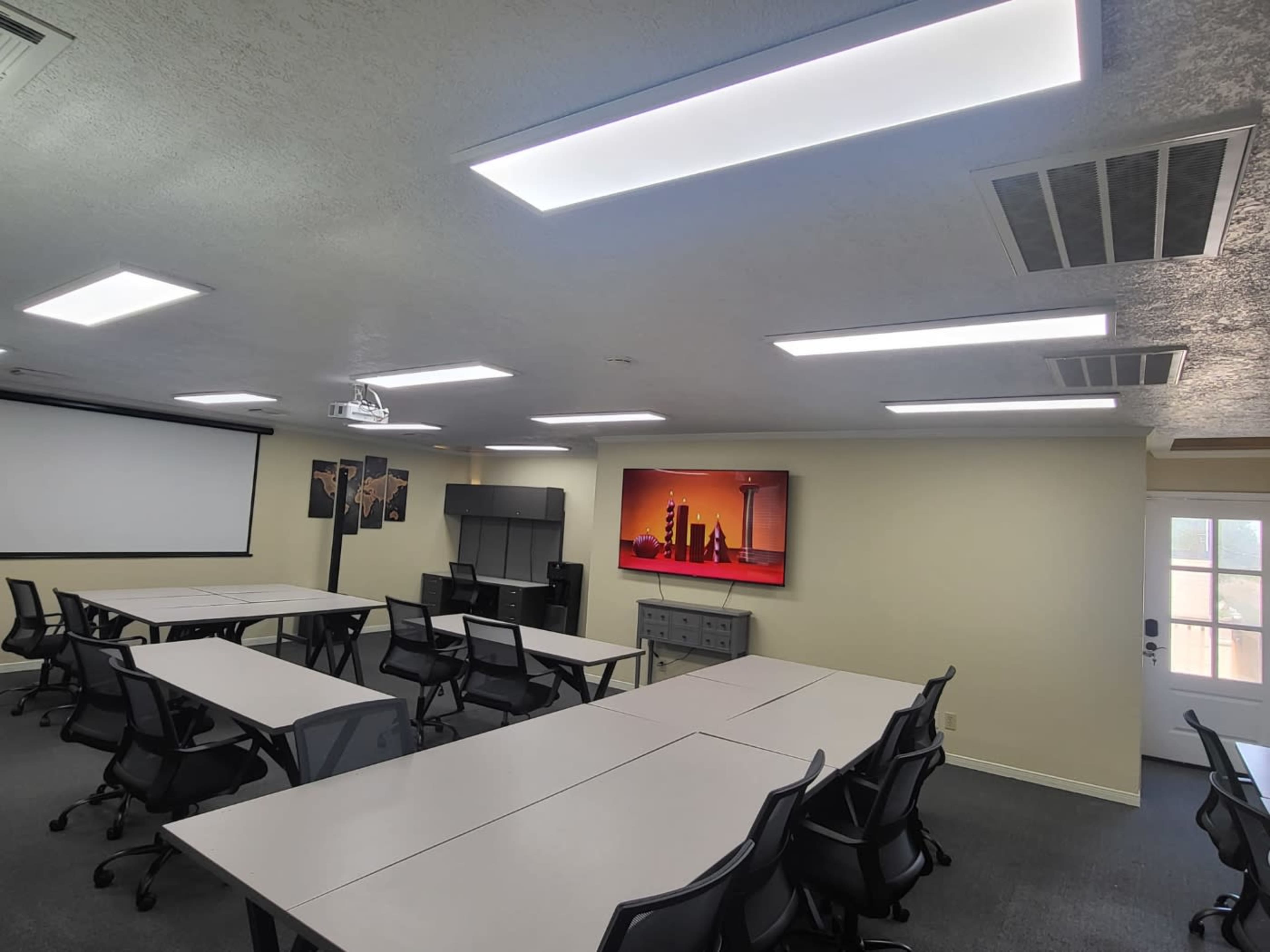 A modern conference room features multiple tables, black chairs, bright overhead lighting, and a large screen displaying a colorful image.