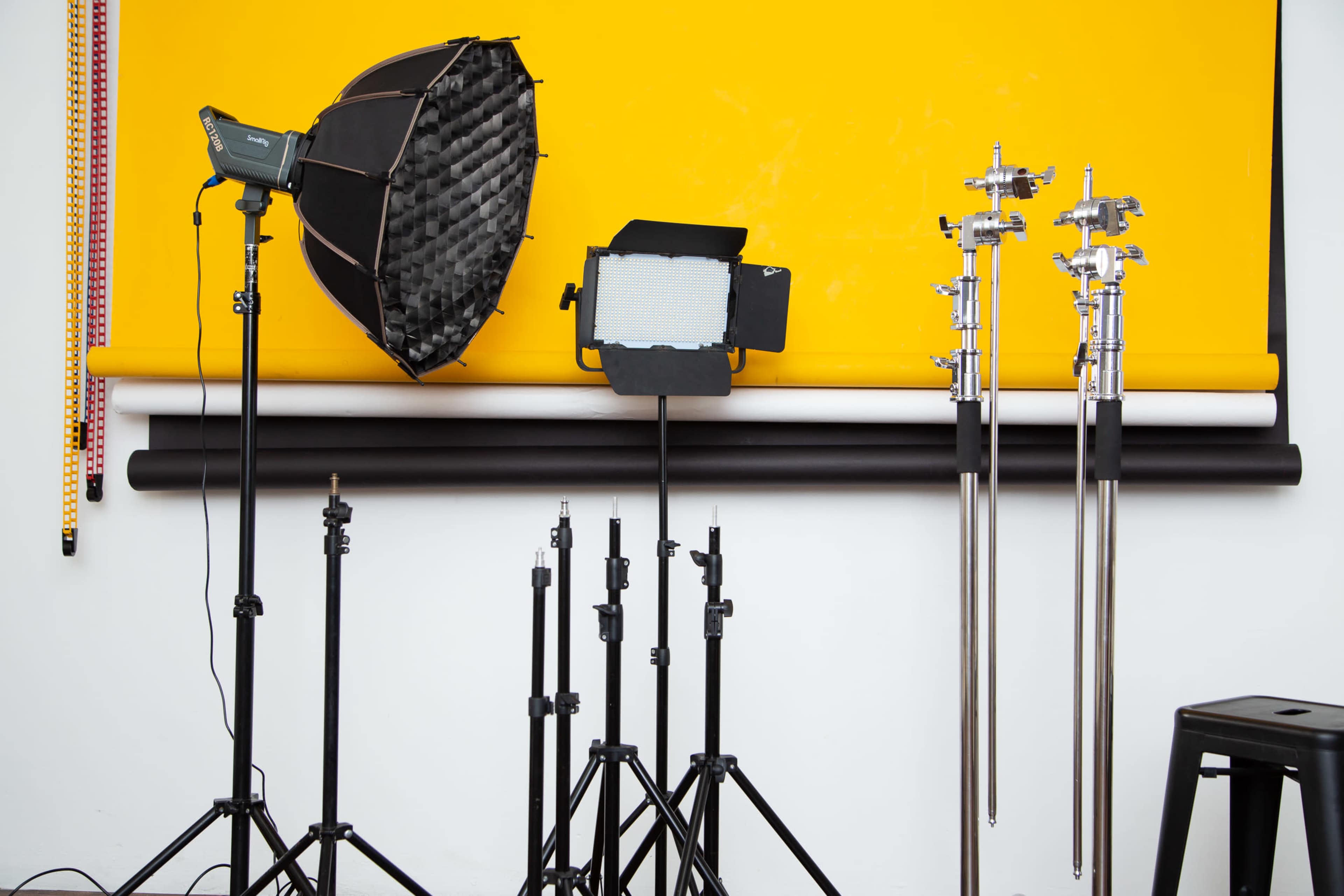 Photography lighting equipment set up against a yellow backdrop, with several light stands and a black stool nearby.