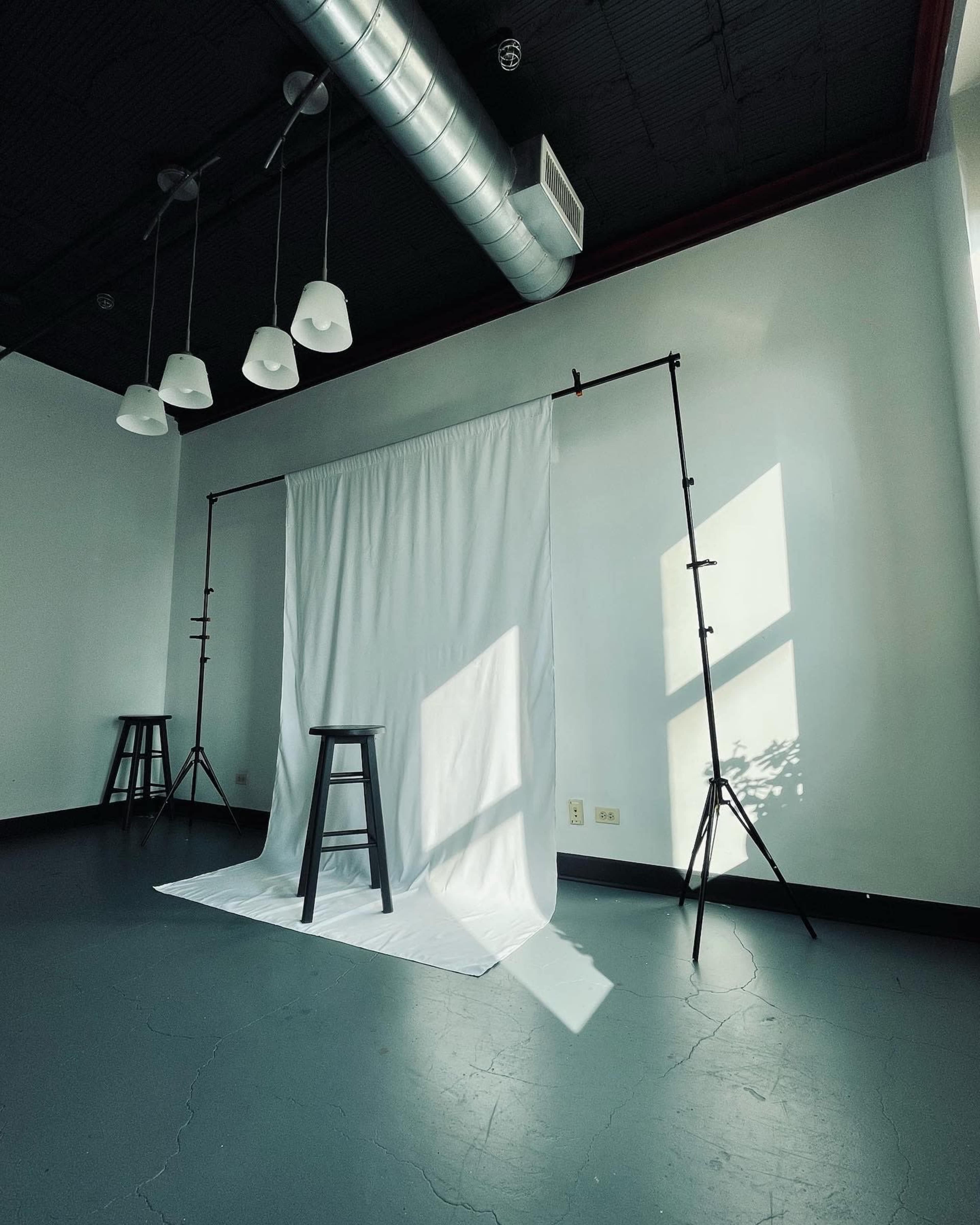A minimalist studio space featuring a white backdrop supported by stands, with a black stool positioned in front, illuminated by natural light.