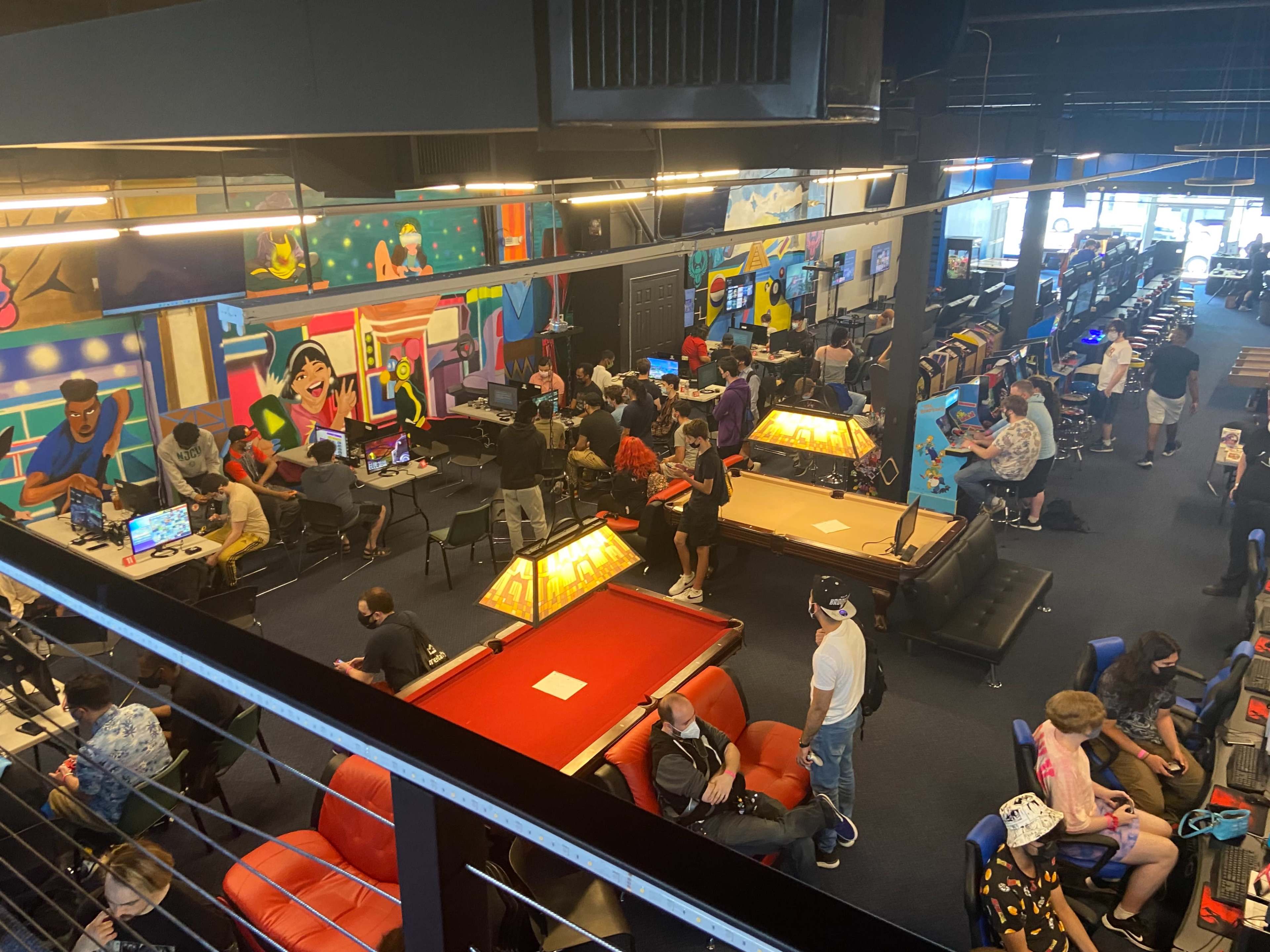 A busy gaming arcade filled with people playing various video games, with colorful murals on the walls and gaming stations set up throughout the space.